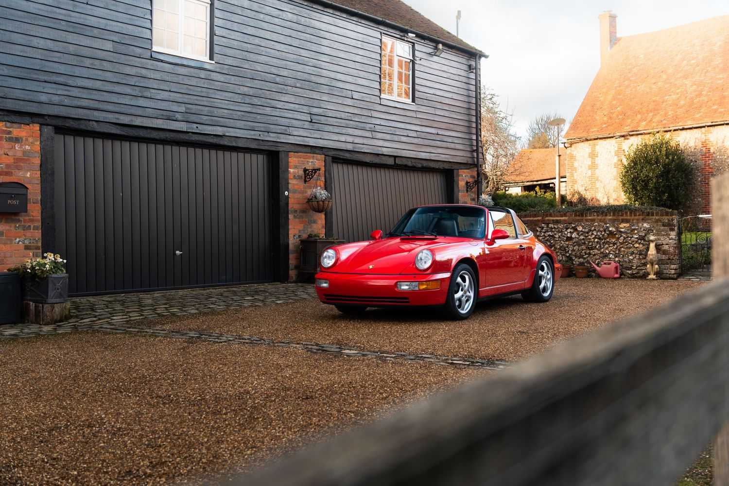 Red Porsche 911 Carrera 2 Targa angled on a gravel driveway beside a dark timber barn and brick building.