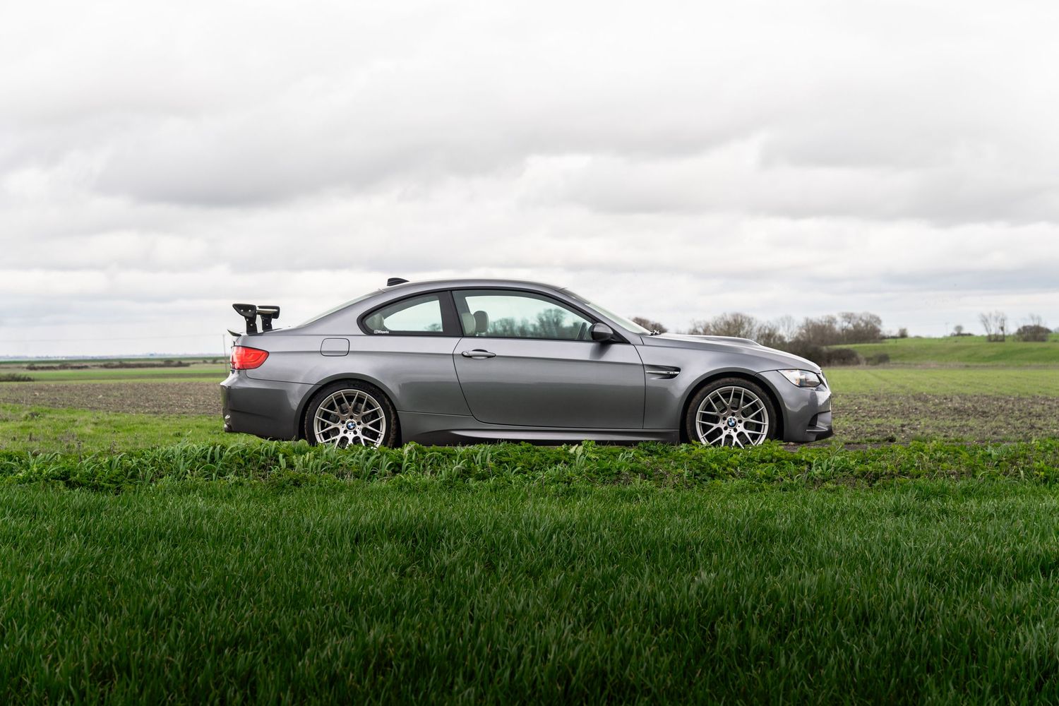 Silver BMW M3 coupe with large rear wing parked on grass field under cloudy sky.