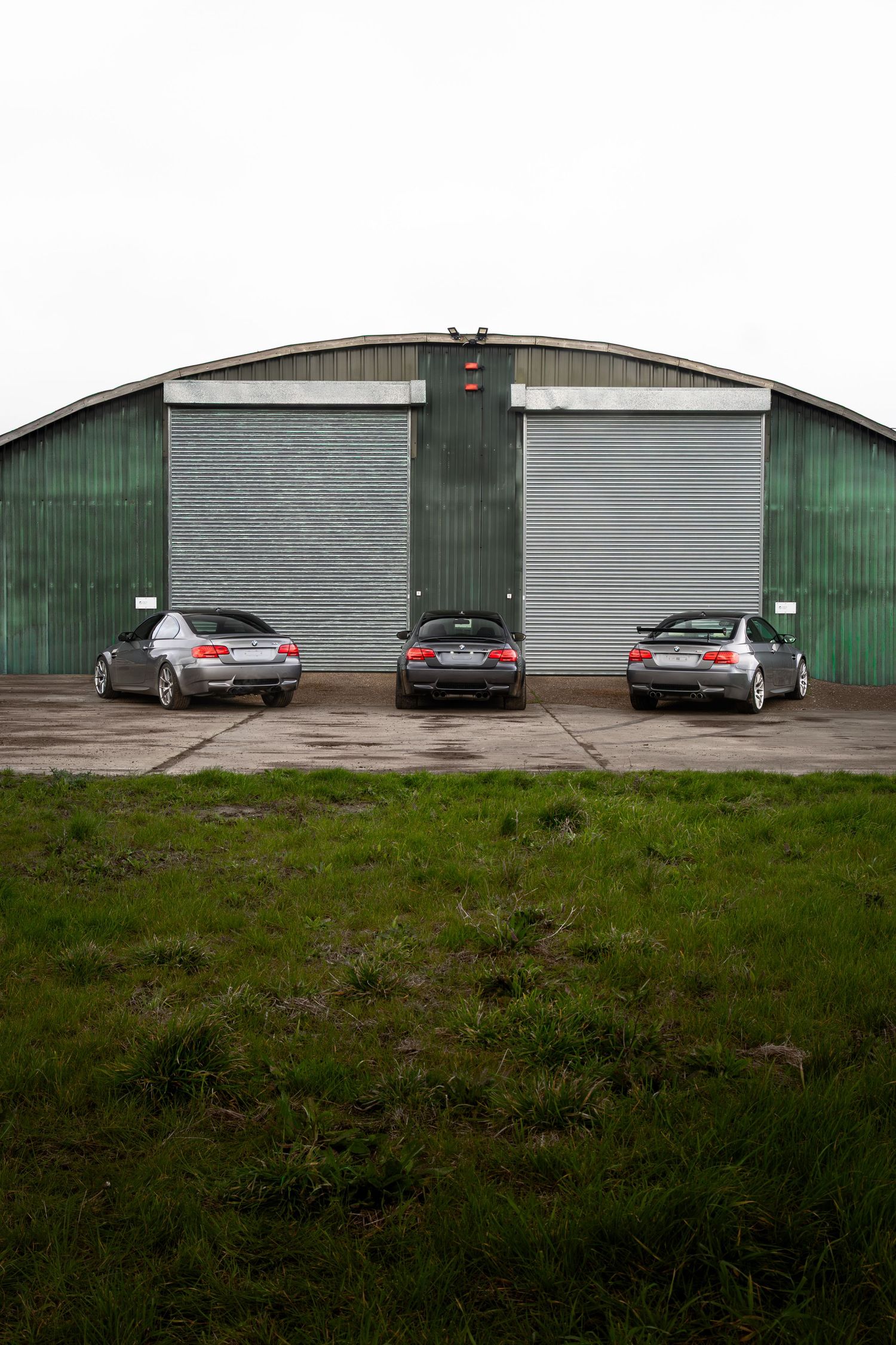 Three BMW M3s parked outside a large green curved-roof hangar building on overcast day.