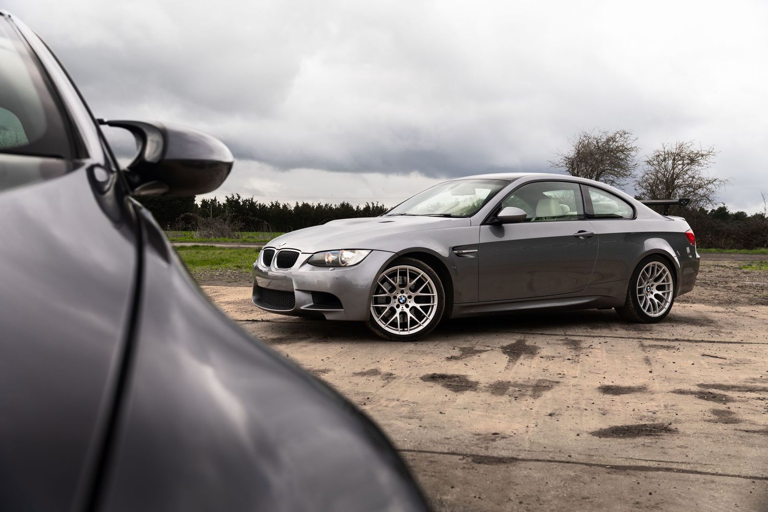 Gray BMW M3 E92 coupe parked on a dirt surface with dramatic cloudy sky and trees in background.