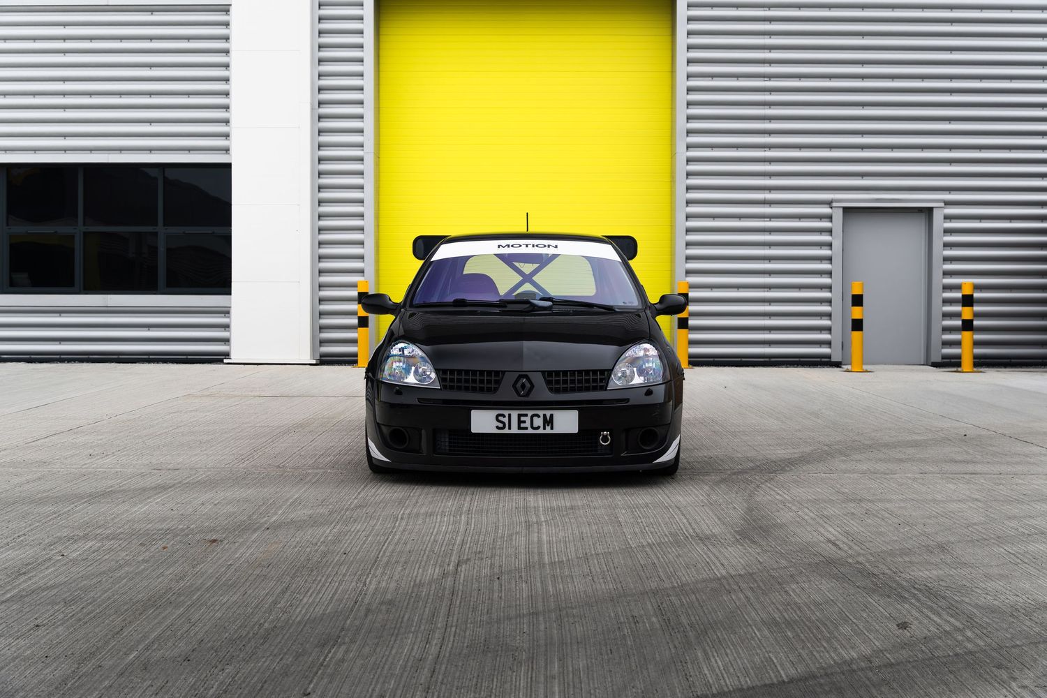 Straight-on front view of a black Renault Clio S1ECM parked in front of yellow garage doors.