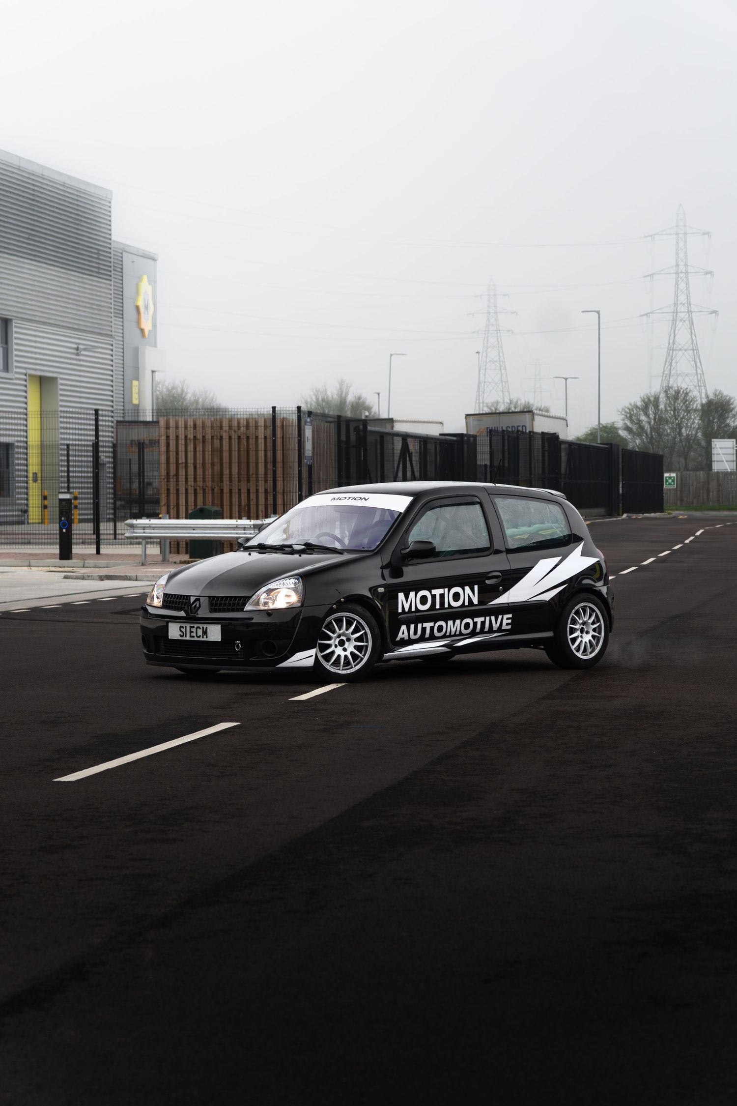 Black Motion Automotive Renault Clio sport on a foggy industrial estate road with power lines visible in background.