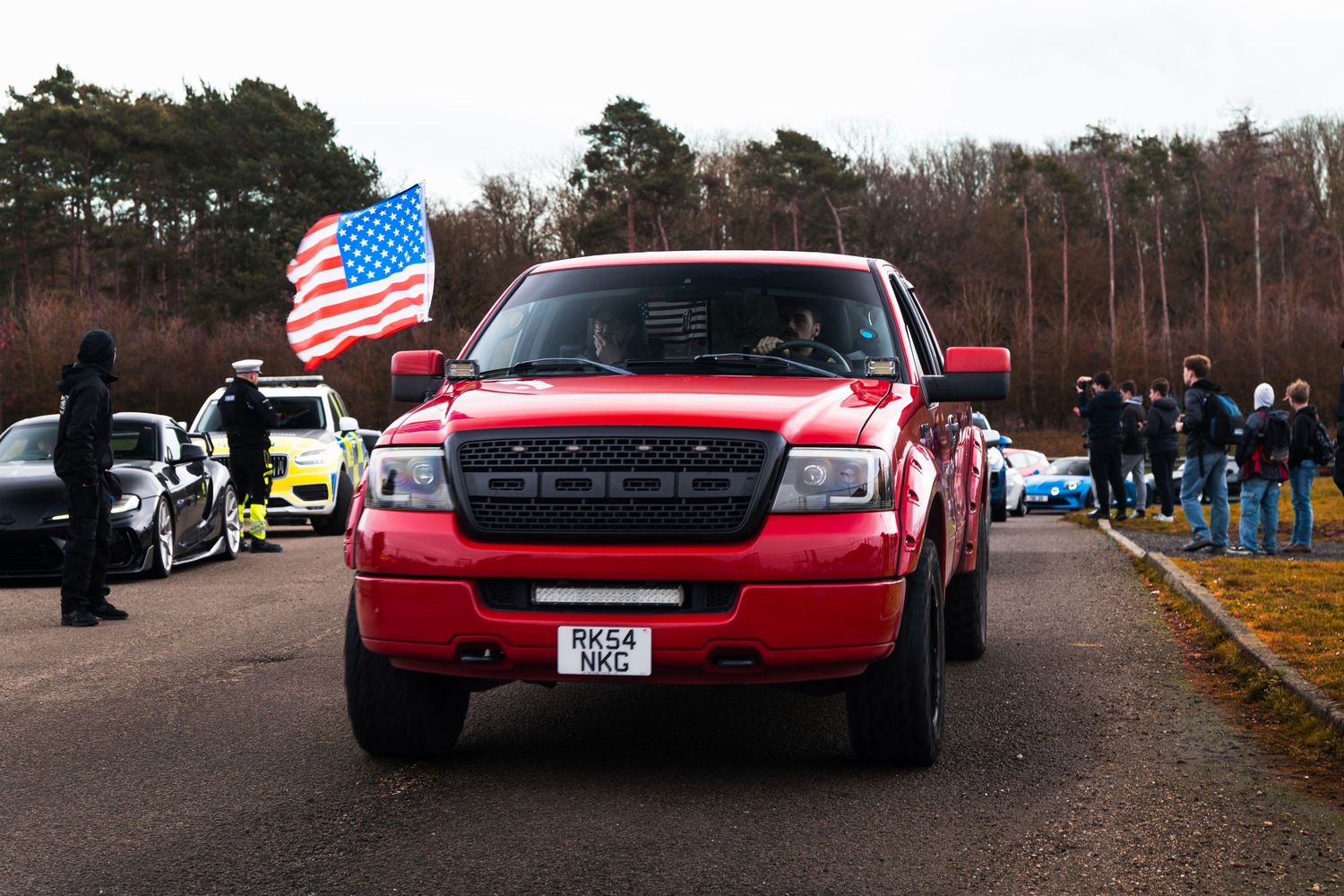 A red Ford pickup truck with an American flag drives through a car meet event on an overcast day.