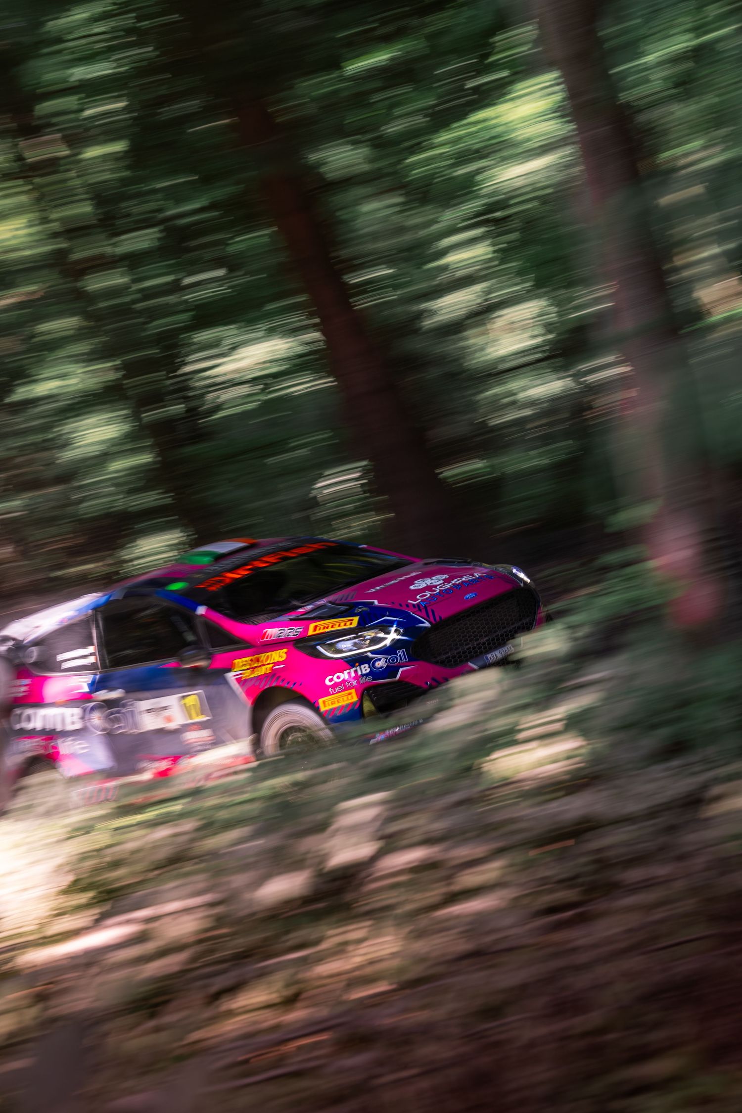 A pink and blue rally car speeds through a forest stage, captured with motion blur emphasizing its high velocity.