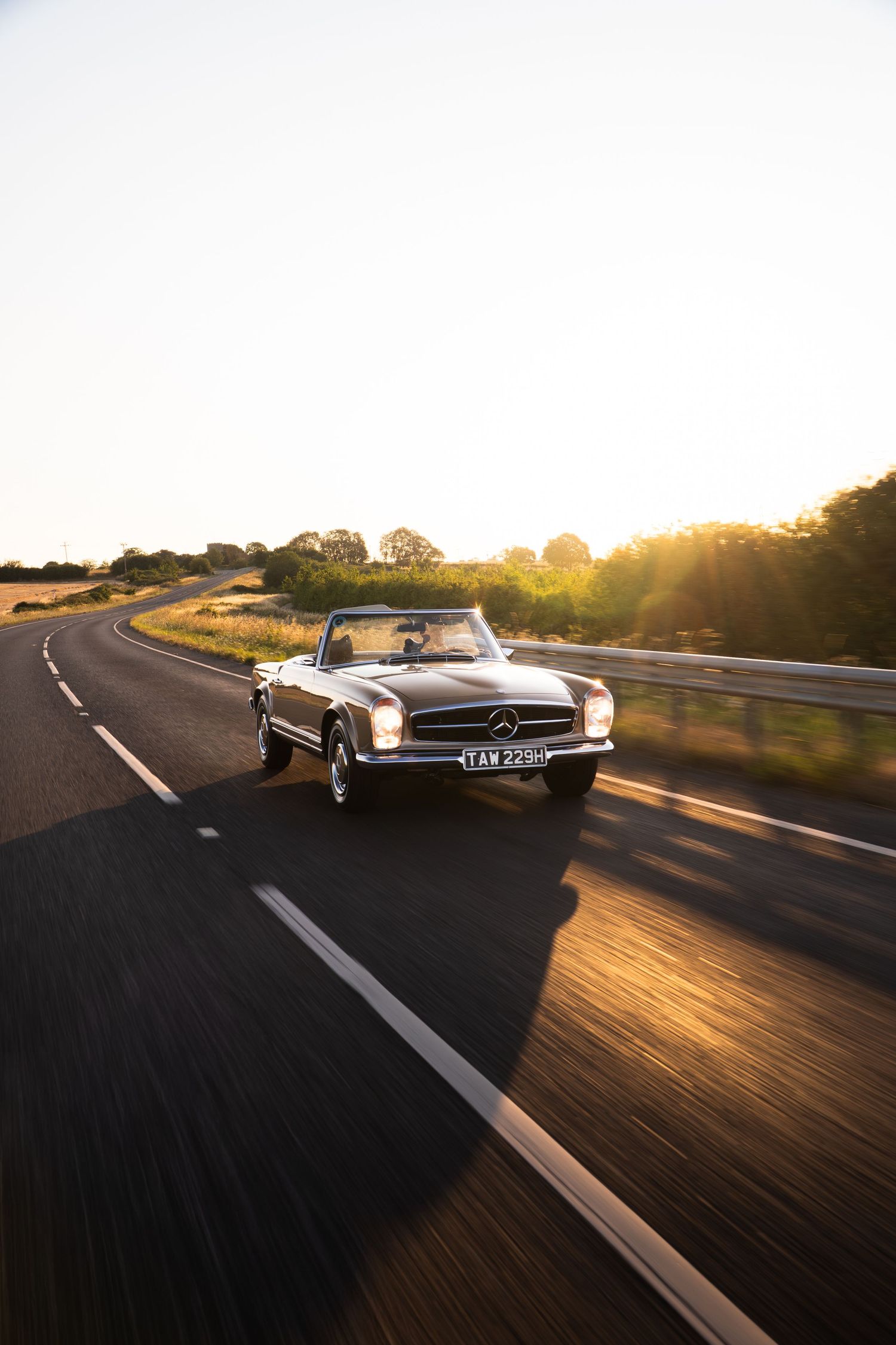 Classic Mercedes-Benz convertible cruising on a sunlit country road at golden hour.