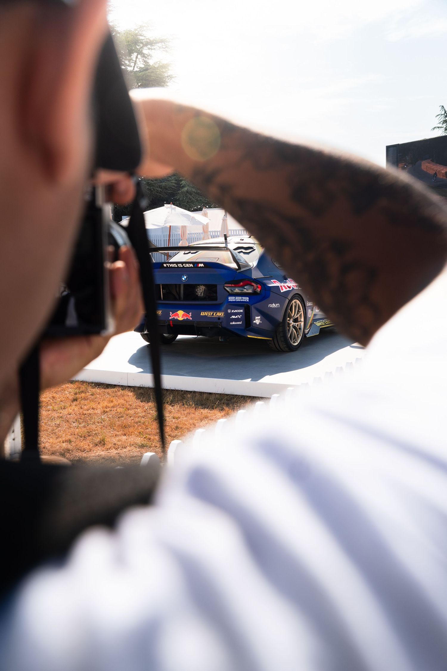 Tattooed photographer capturing a blue Red Bull racing car at an outdoor motorsport event on a sunny day.