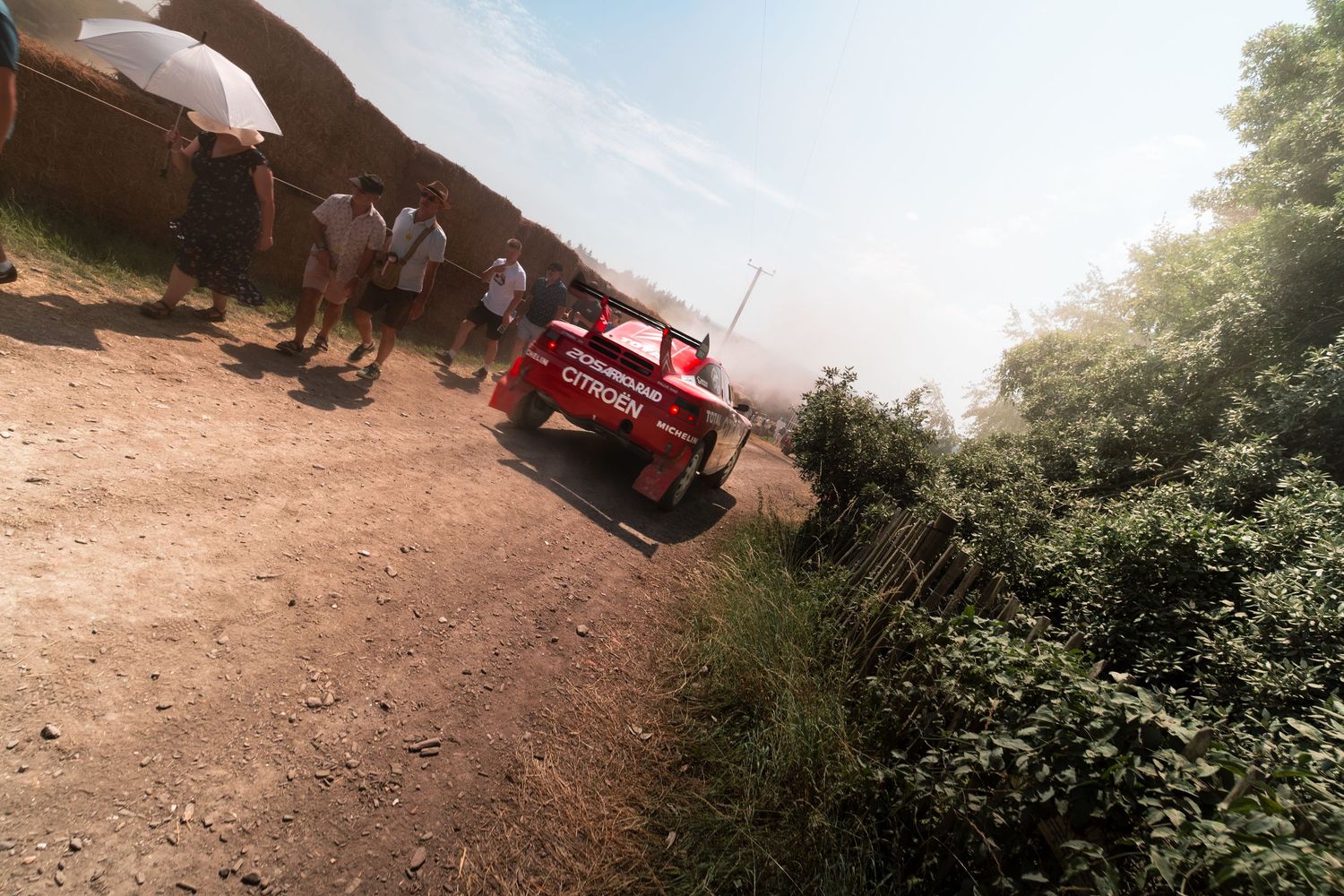 Red Citroën rally car on a dusty dirt road with spectators watching during an outdoor rally event.