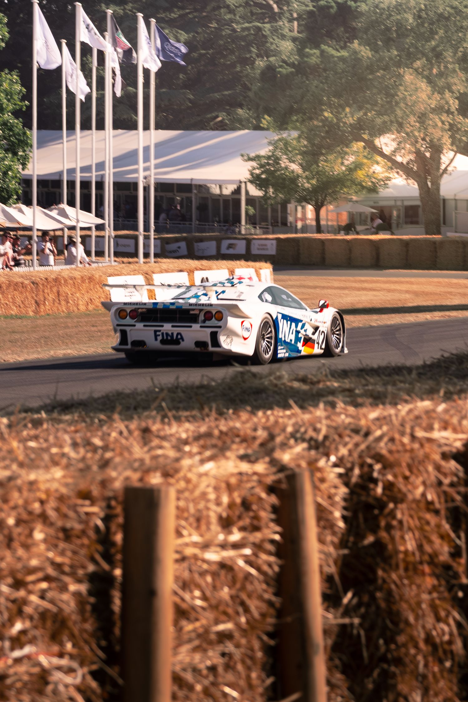 White GT racing car navigates a hay bale lined track at Goodwood Festival of Speed with flags in background.