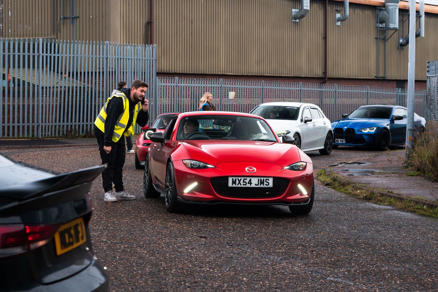 A man in a hi-vis vest inspects a red Mazda MX-5 in a car meet parking lot.
