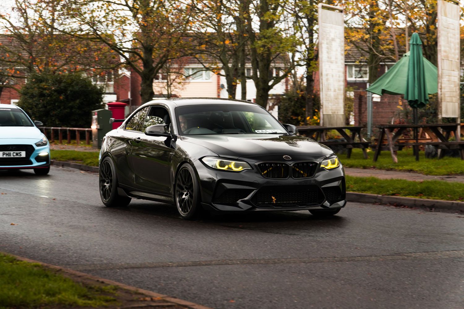 Dark grey BMW M2 with yellow angel eyes driving on wet UK residential street in autumn.