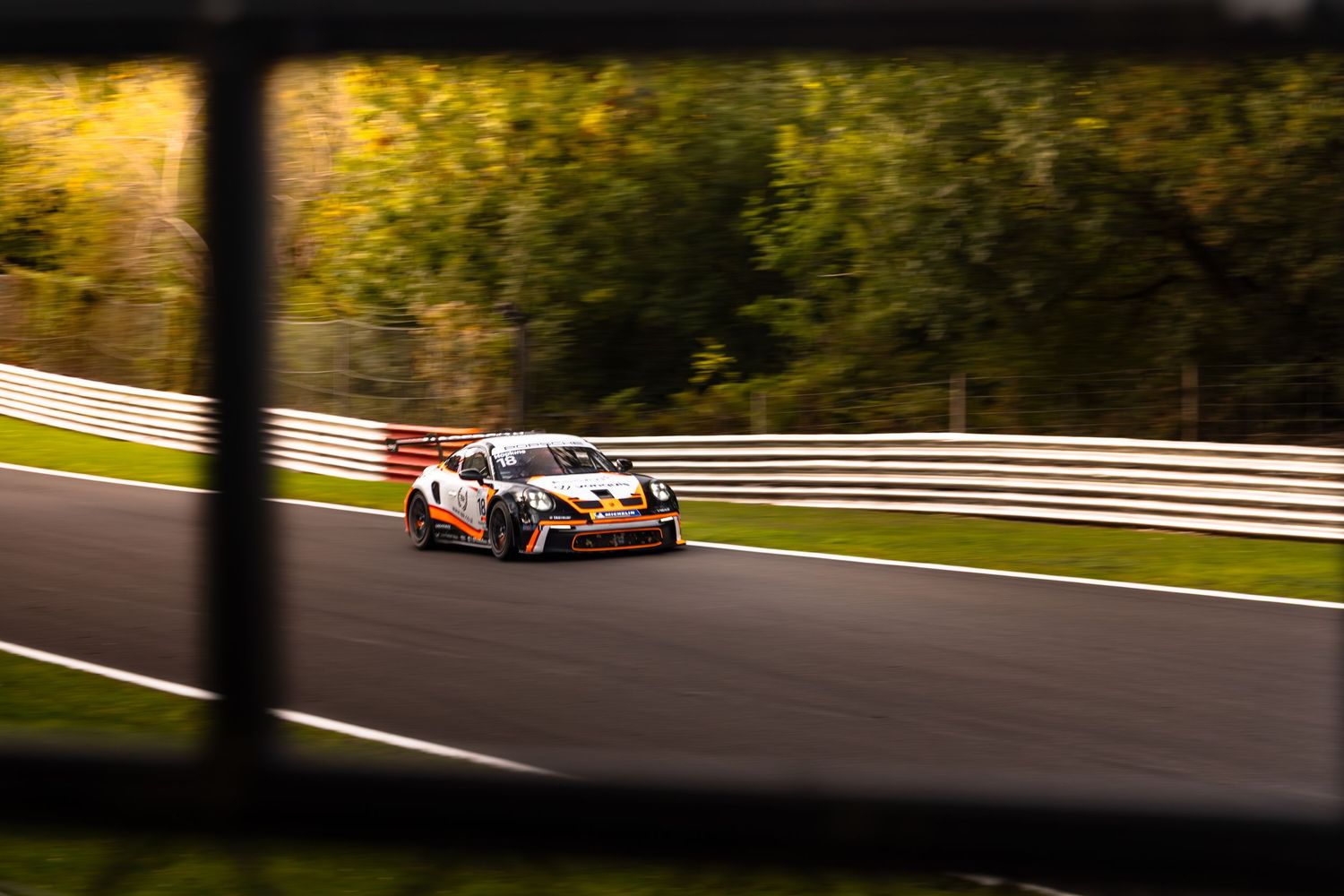 Orange and white Porsche GT3 race car navigating a corner on a circuit, shot through a fence.