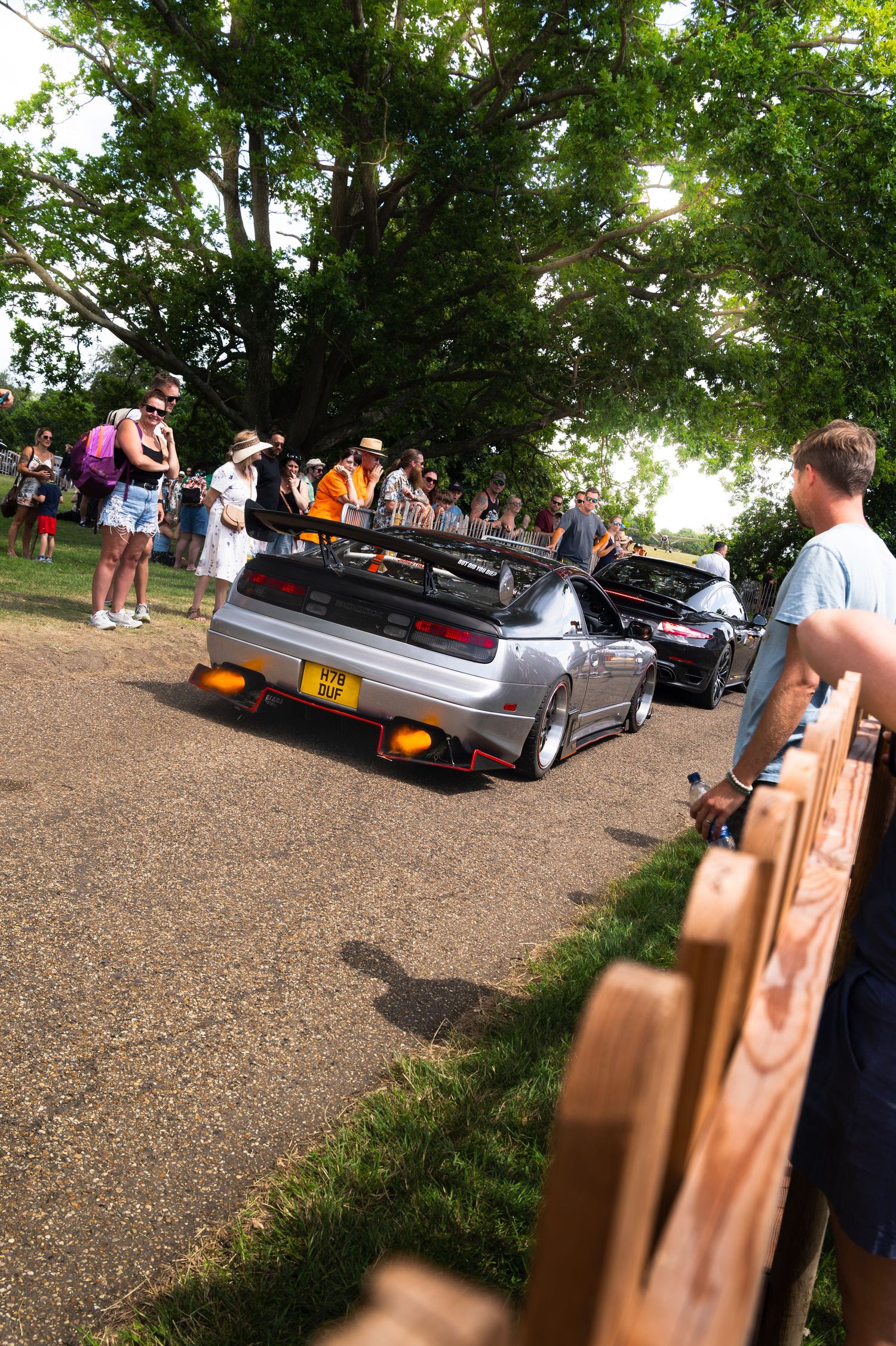 Silver modified car with large spoiler at outdoor car show, crowd of spectators gathered under green trees on sunny day.