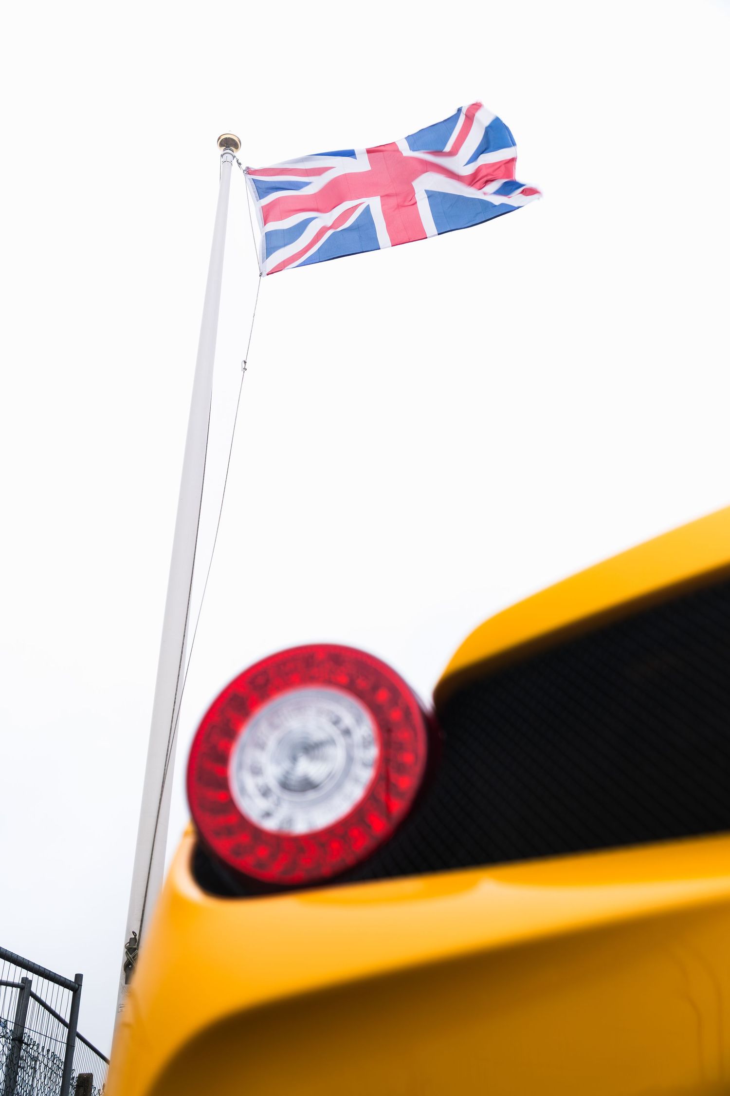 Yellow Ferrari with red tail light detail, Union Jack flag flying on pole against white sky at UK motorsport event.