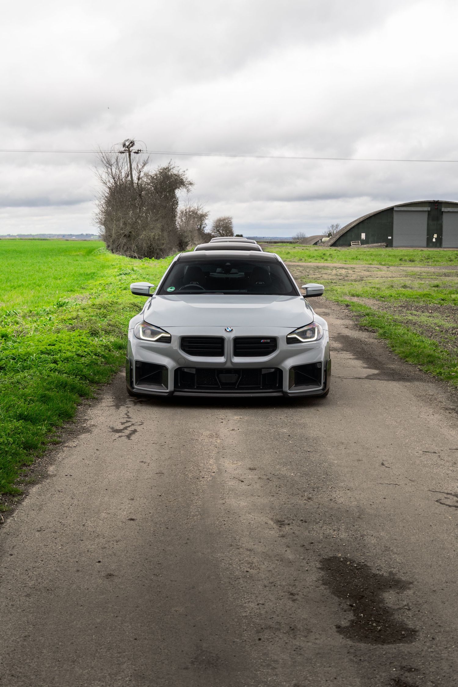 Front view of a white BMW M2 Competition on a dirt road between green fields and a barn.