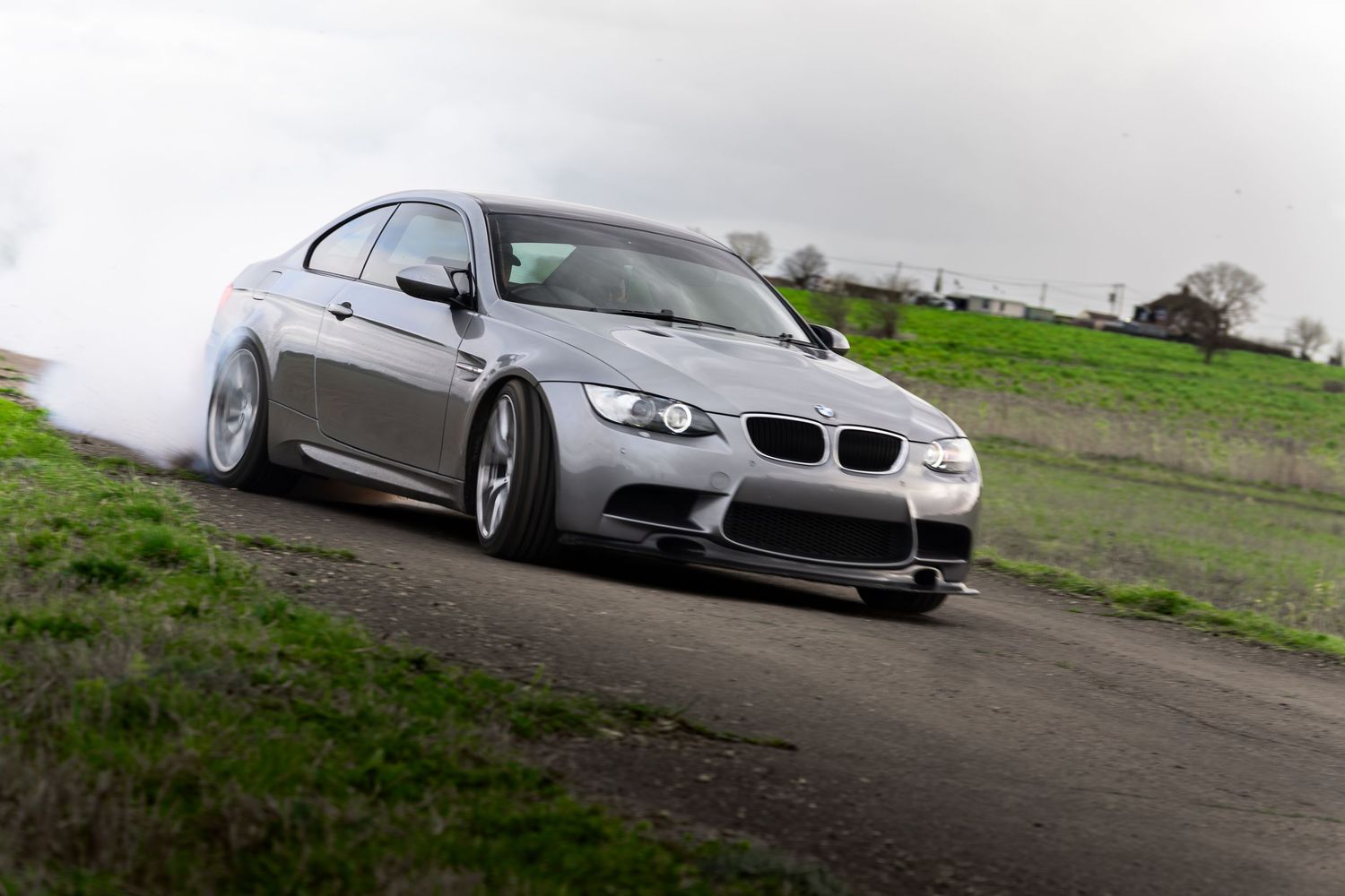 Silver BMW M3 E92 drifting aggressively with heavy tire smoke on a countryside road.