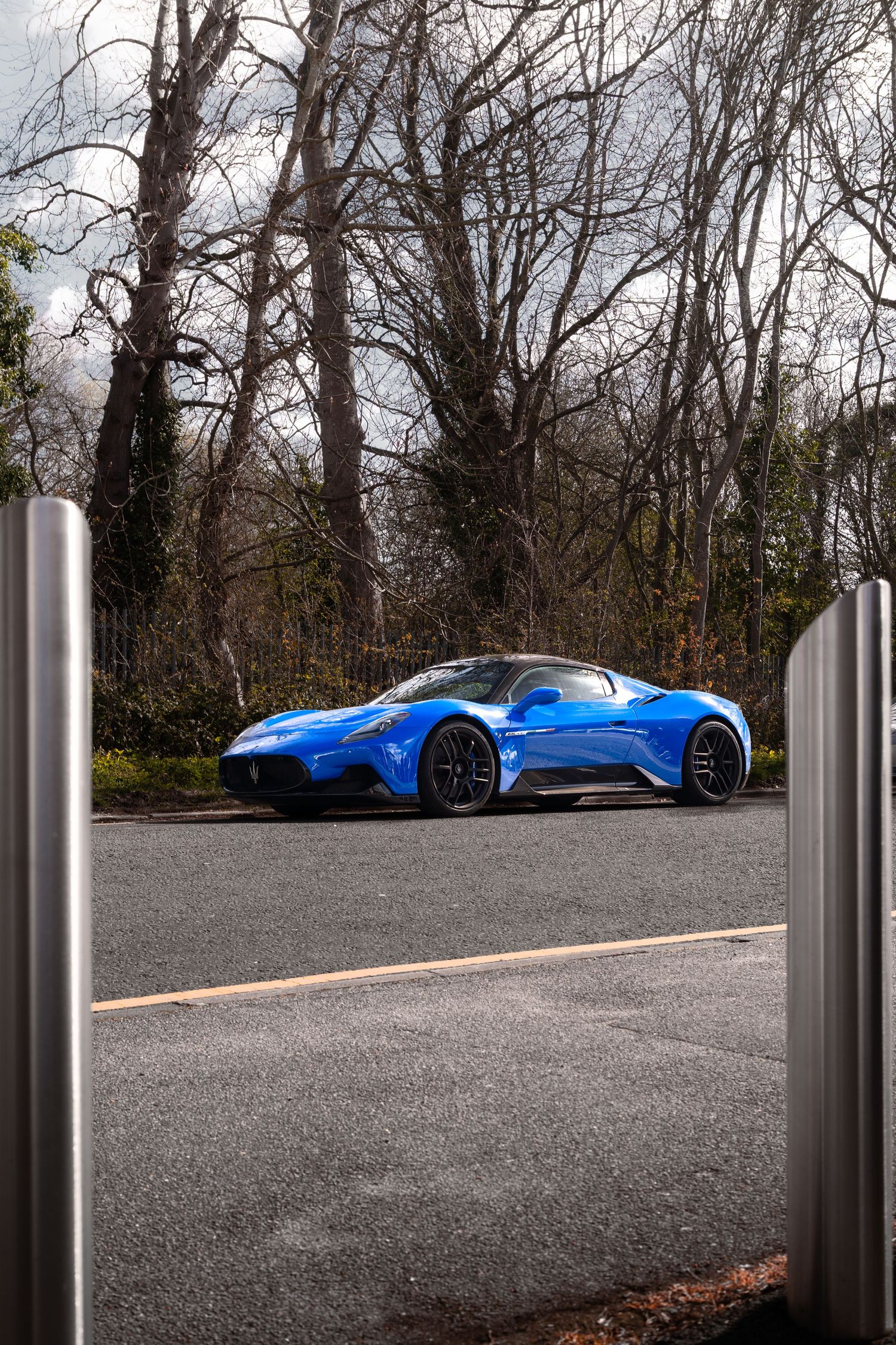 Blue supercar parked on a wet road surrounded by bare trees on a cloudy day.