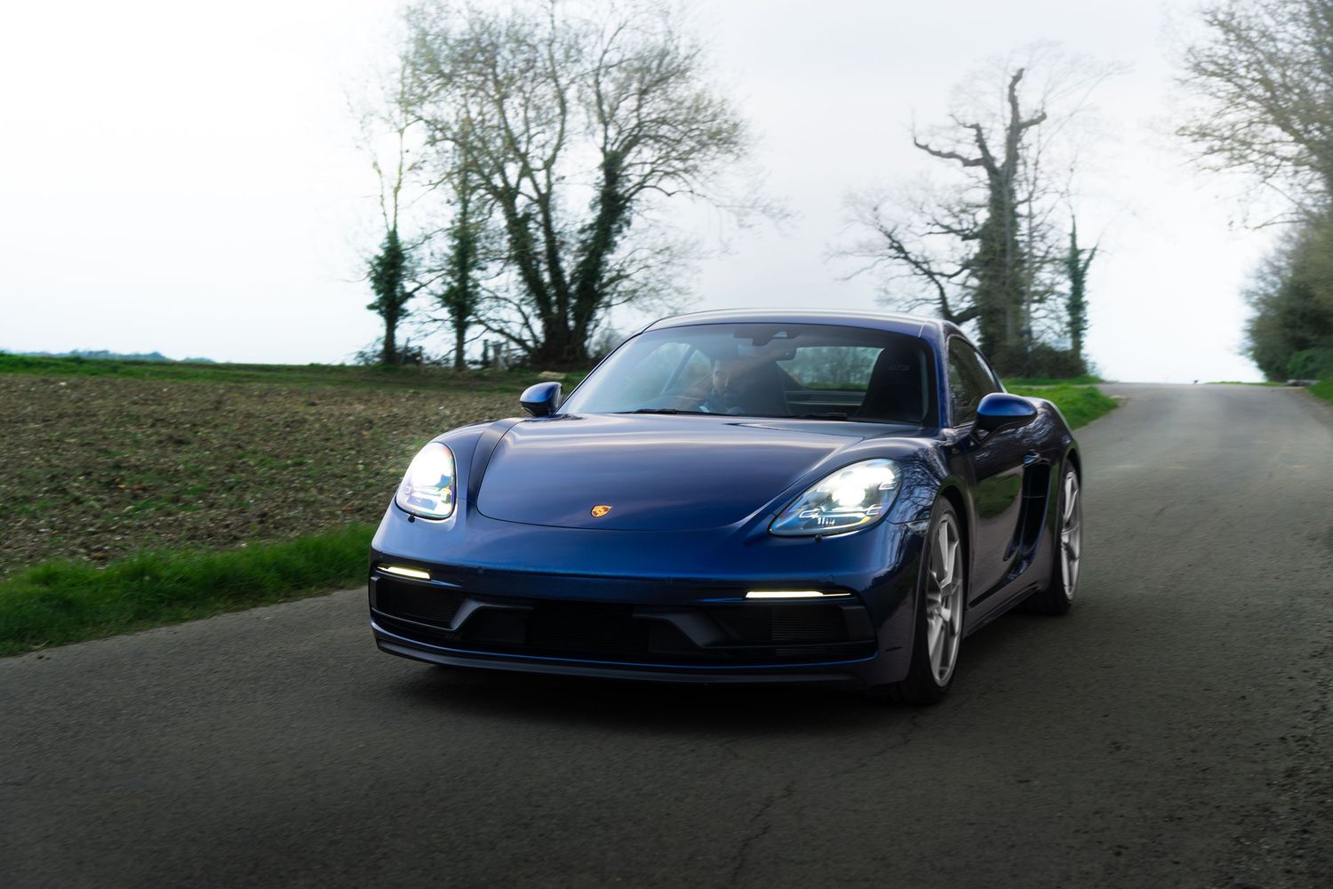 Blue Porsche 718 Cayman GTS driving on a rural road surrounded by bare winter trees.