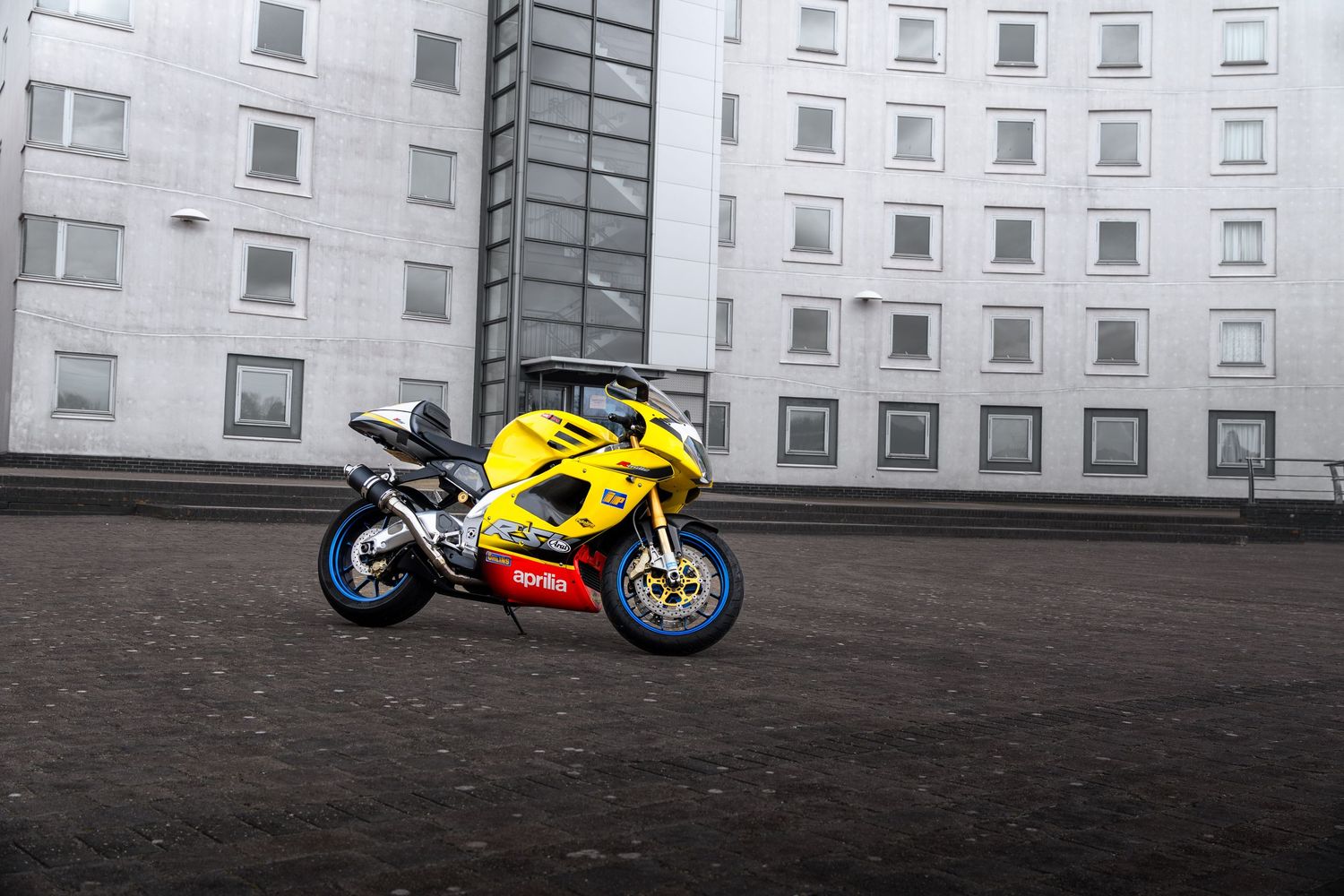 Yellow Aprilia RSV superbike parked on dark cobblestones beside a large grey modern building.