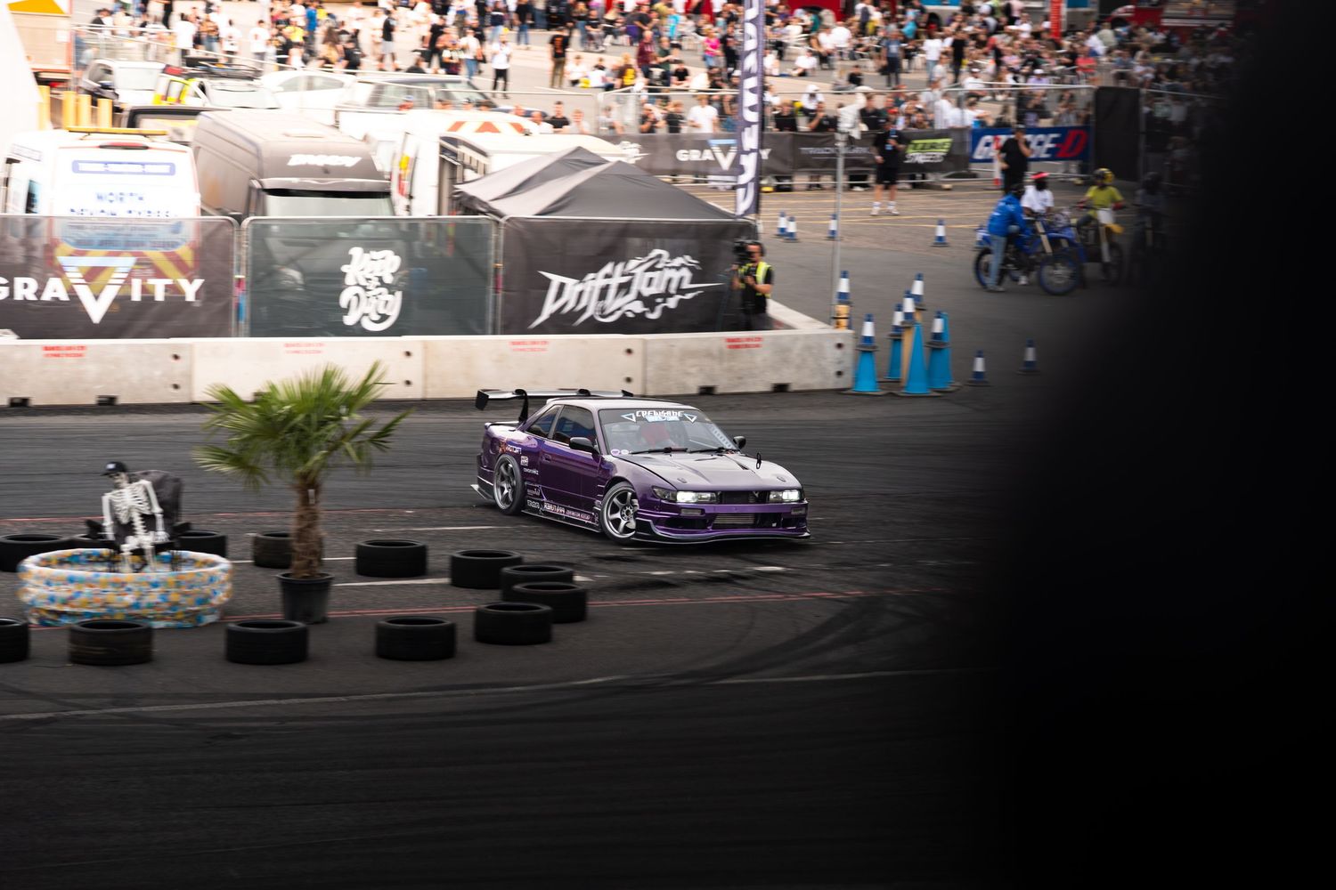 A purple modified drift car navigates a night circuit surrounded by tires and palm trees at a Drift King event.