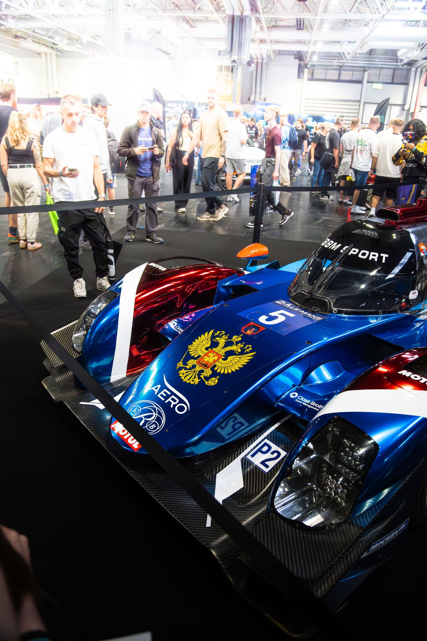 Blue LMP racing car on display at motorsport expo with crowds of visitors in background.