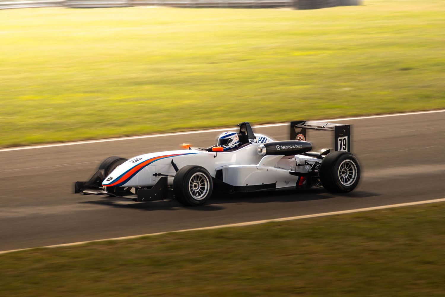 White Formula racing car with orange stripe speeds along a sunlit race circuit track.