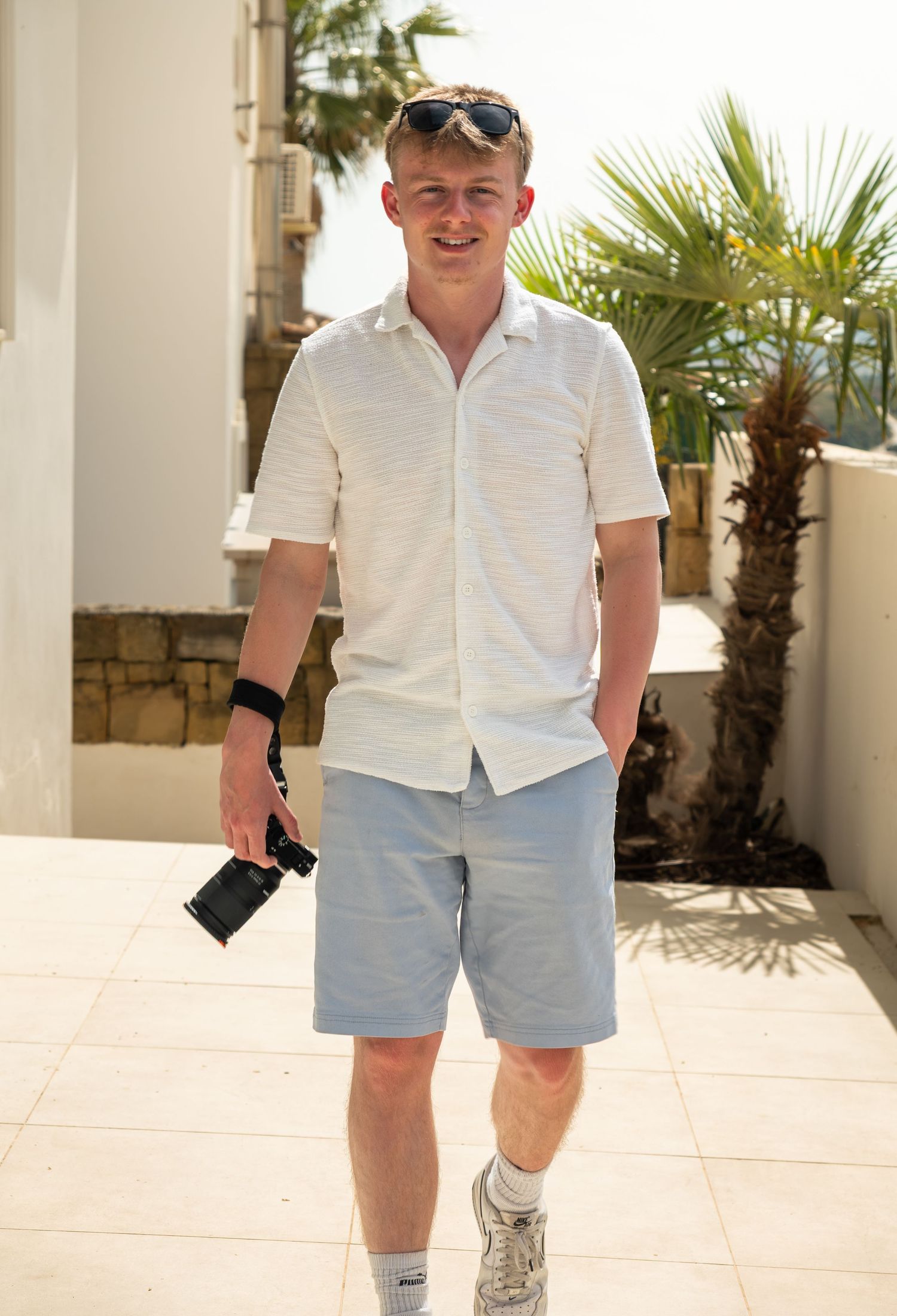 Smiling young man with sunglasses holding a camera walking on a bright sunny outdoor terrace.