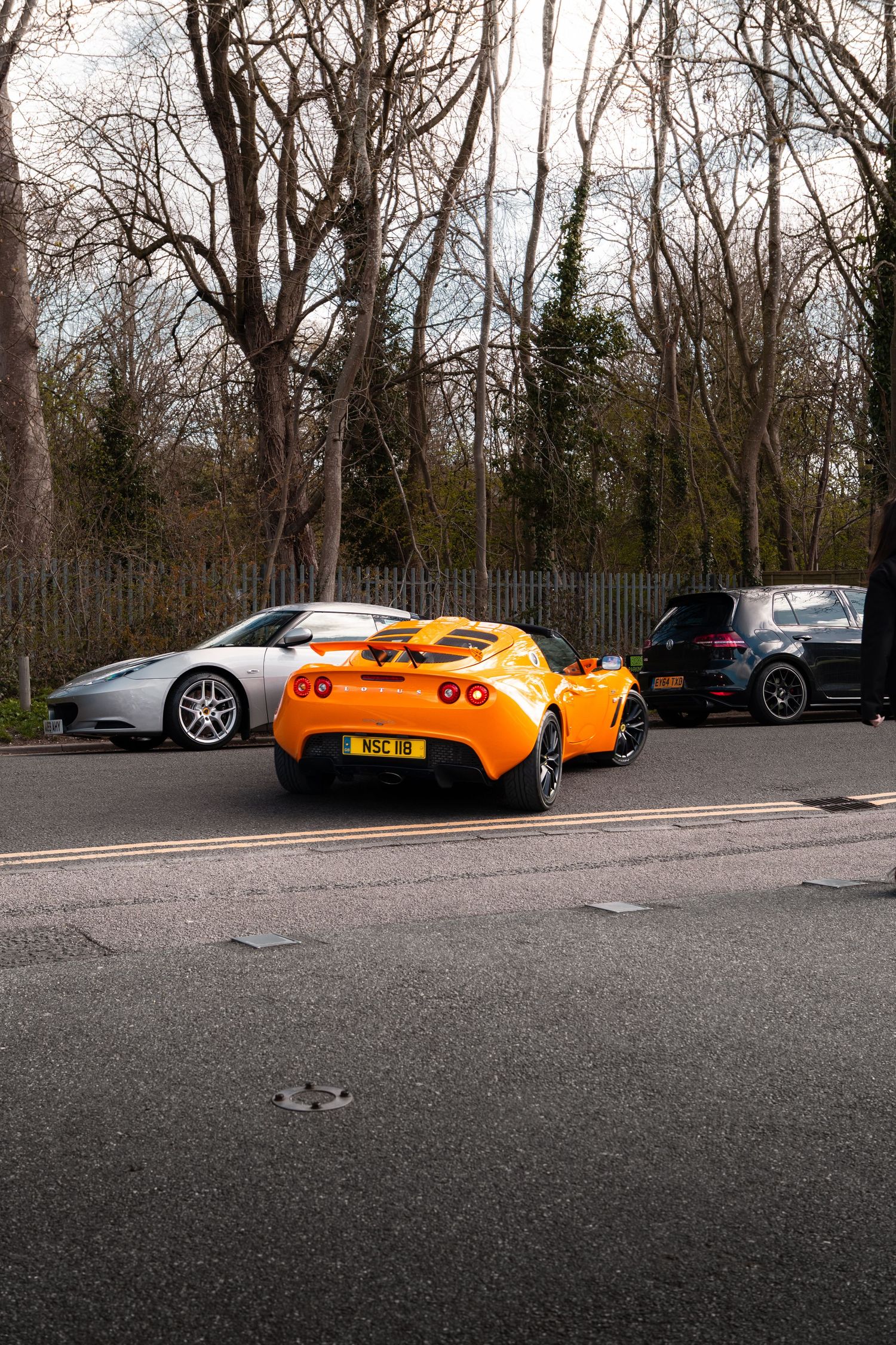 Orange Lotus Elise and silver sports car parked on a road surrounded by bare winter trees.