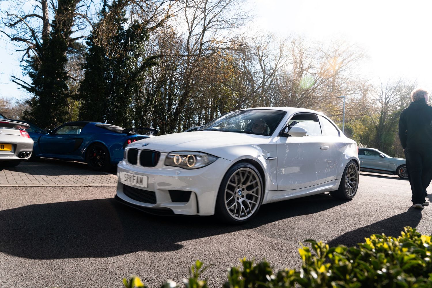 White BMW 1M Coupe with aftermarket wheels parked in a sunny lot with trees in the background.