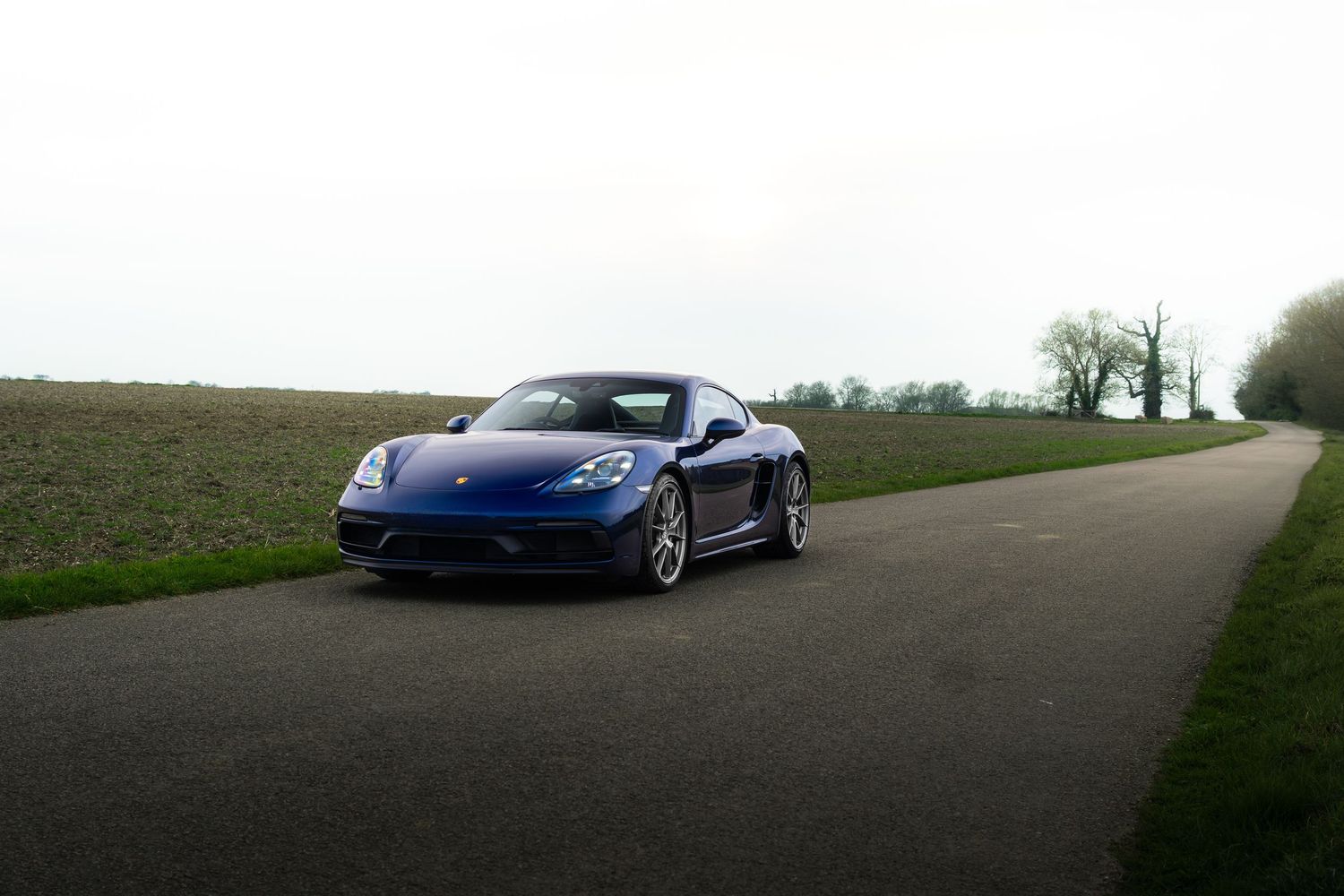 Blue Porsche 718 Cayman driving on a country road surrounded by green fields and bare trees.