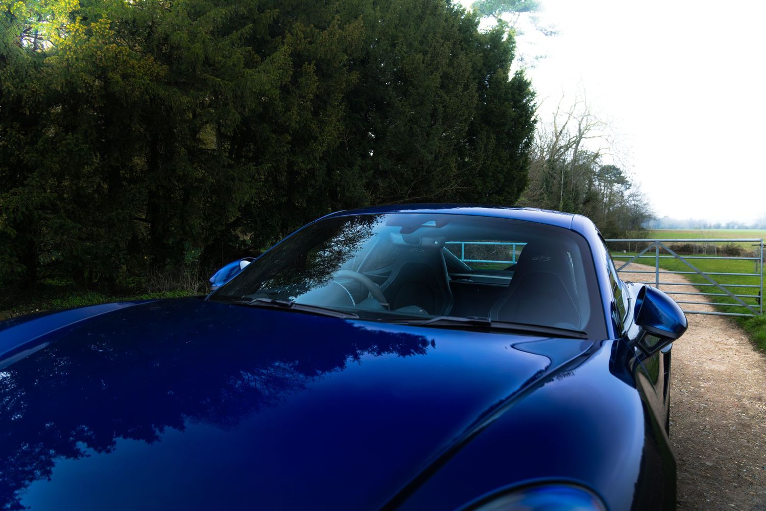 Top view of blue Porsche 718 Cayman GTS roof and windscreen reflecting trees on a rural driveway.