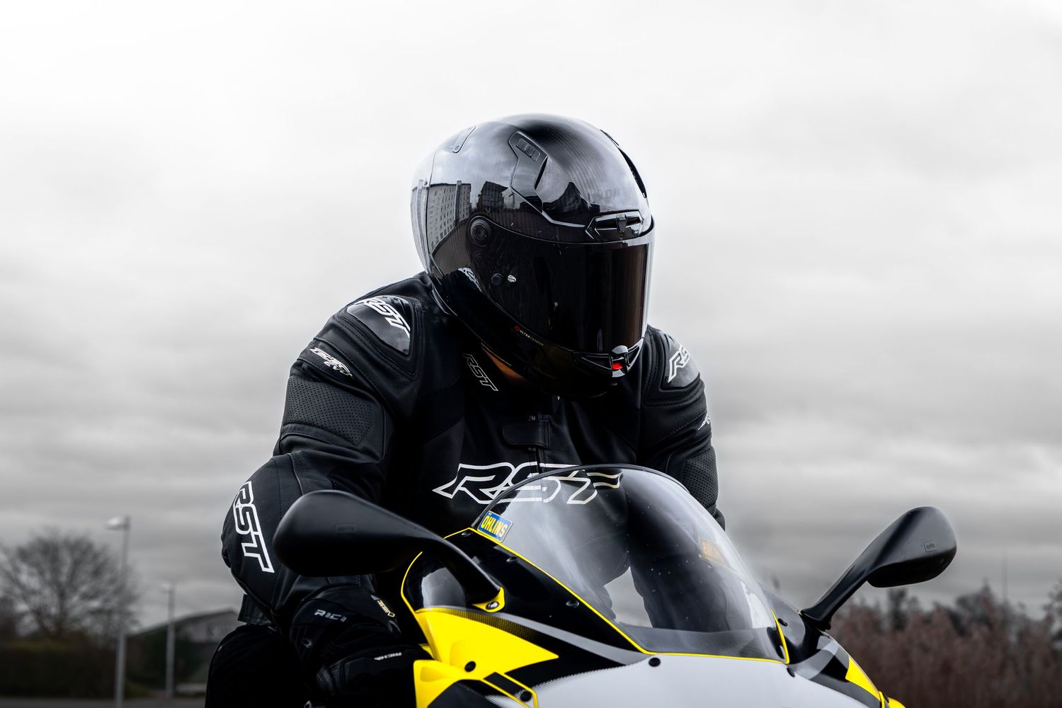 Close-up of a motorcyclist in RST black leather jacket and dark helmet on a yellow sportbike.