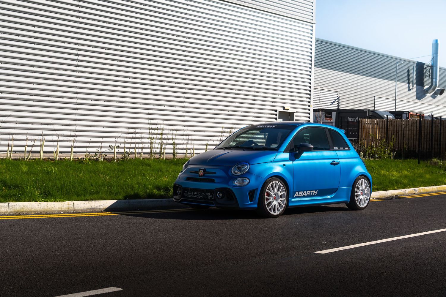 Side profile of a lowered blue Abarth 500 with white alloy wheels parked beside a corrugated metal building.
