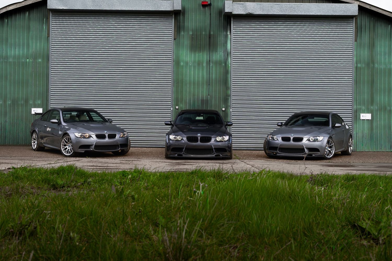 Three grey BMW M3 cars displayed in front of green garage doors with grass foreground.
