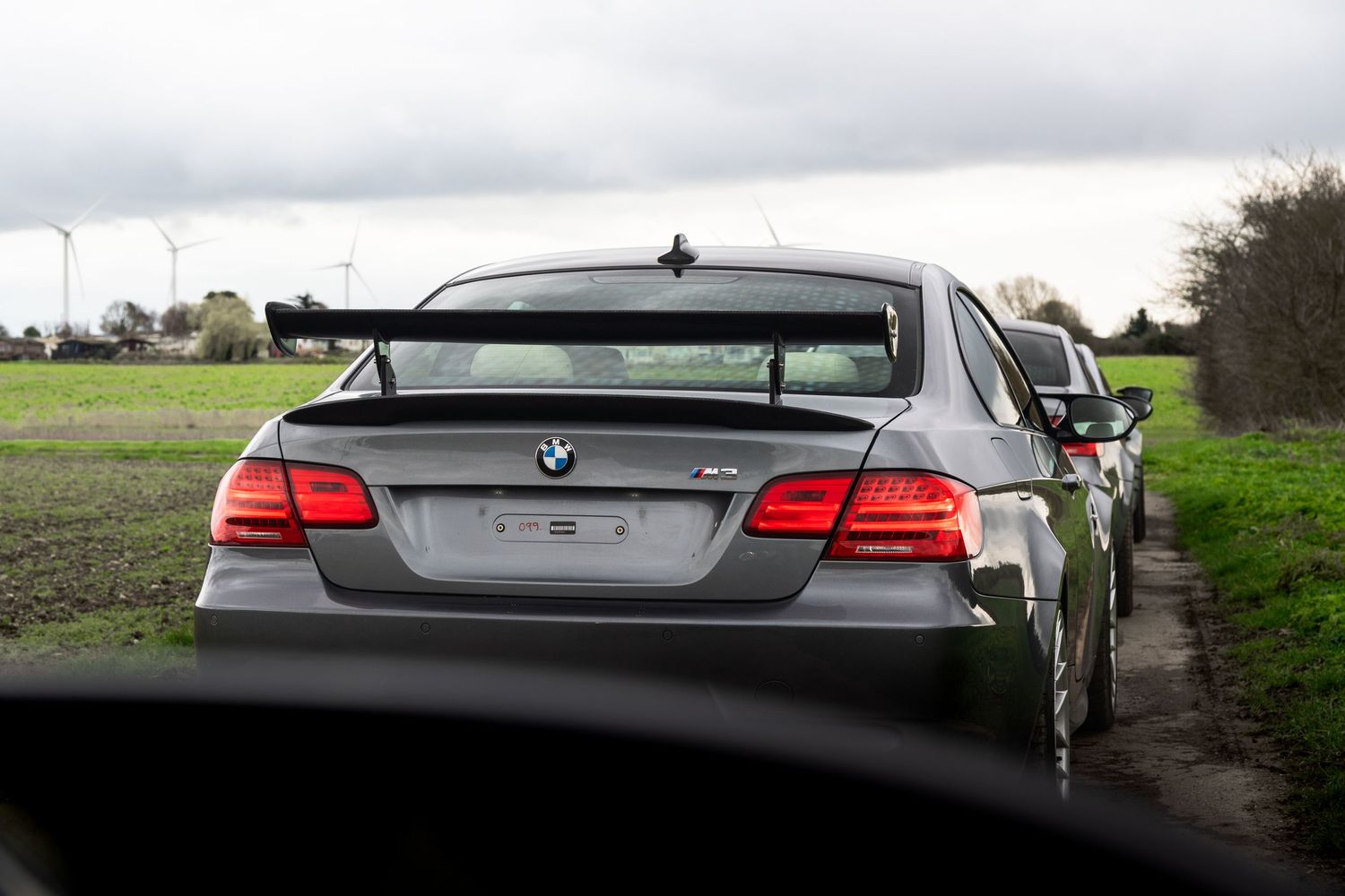 Rear view of silver BMW M3 coupe with spoiler parked in green countryside setting.