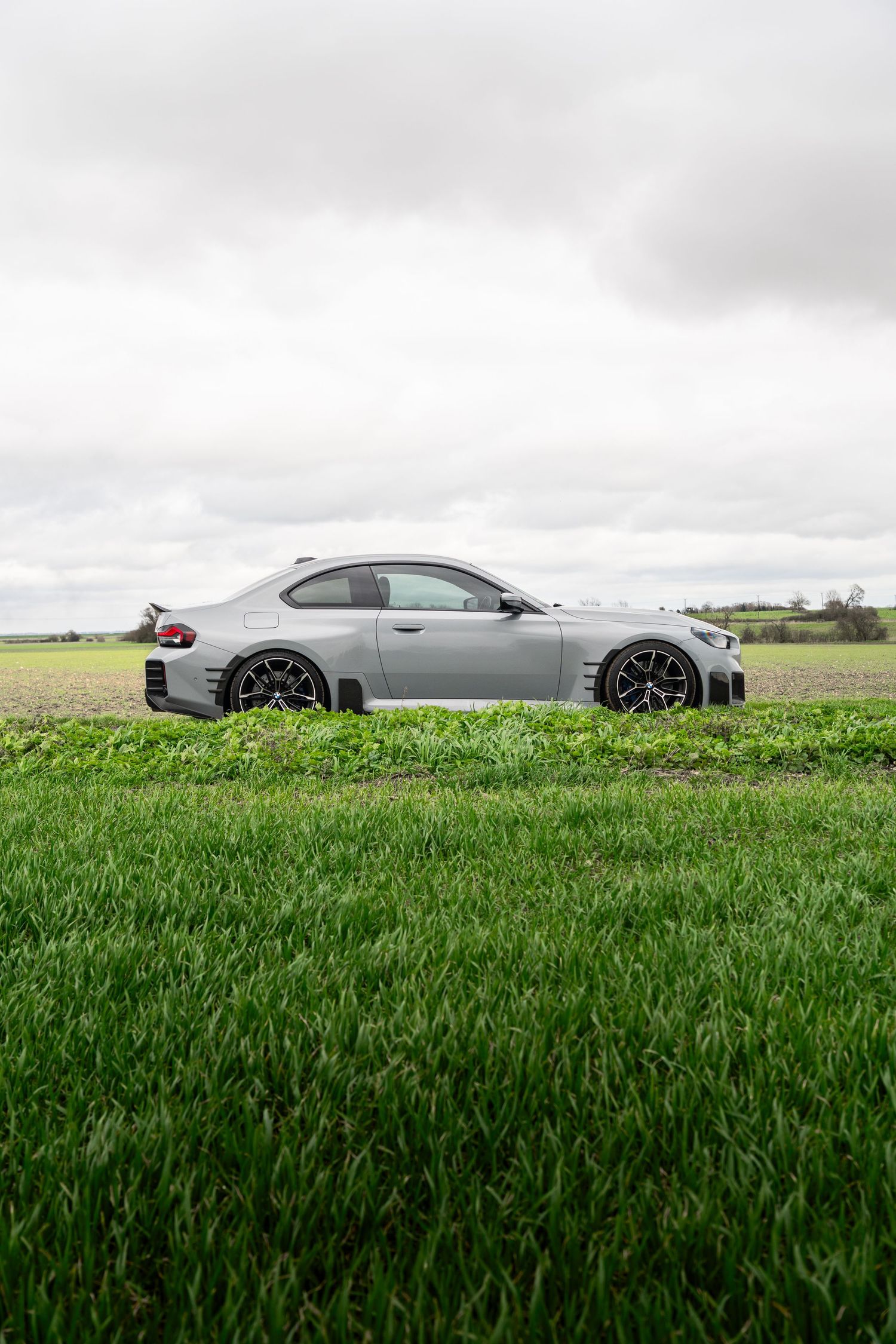 Side profile of a matte gray BMW M2 coupe on a country road surrounded by green fields.
