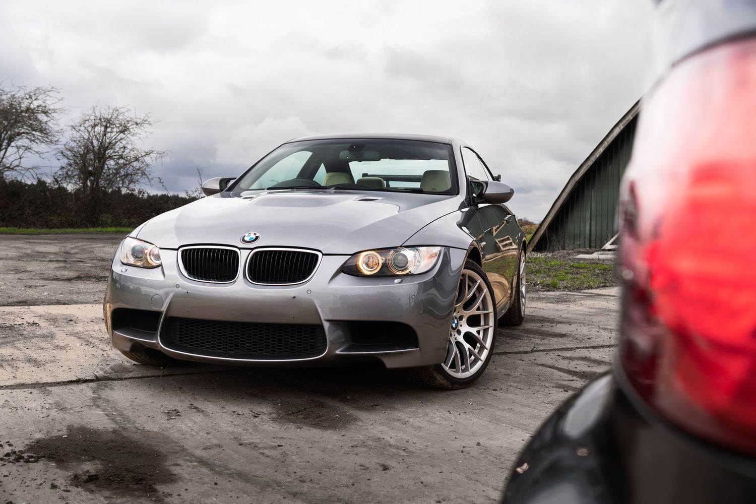Front three-quarter view of a silver BMW M3 coupe on an open tarmac area outdoors.