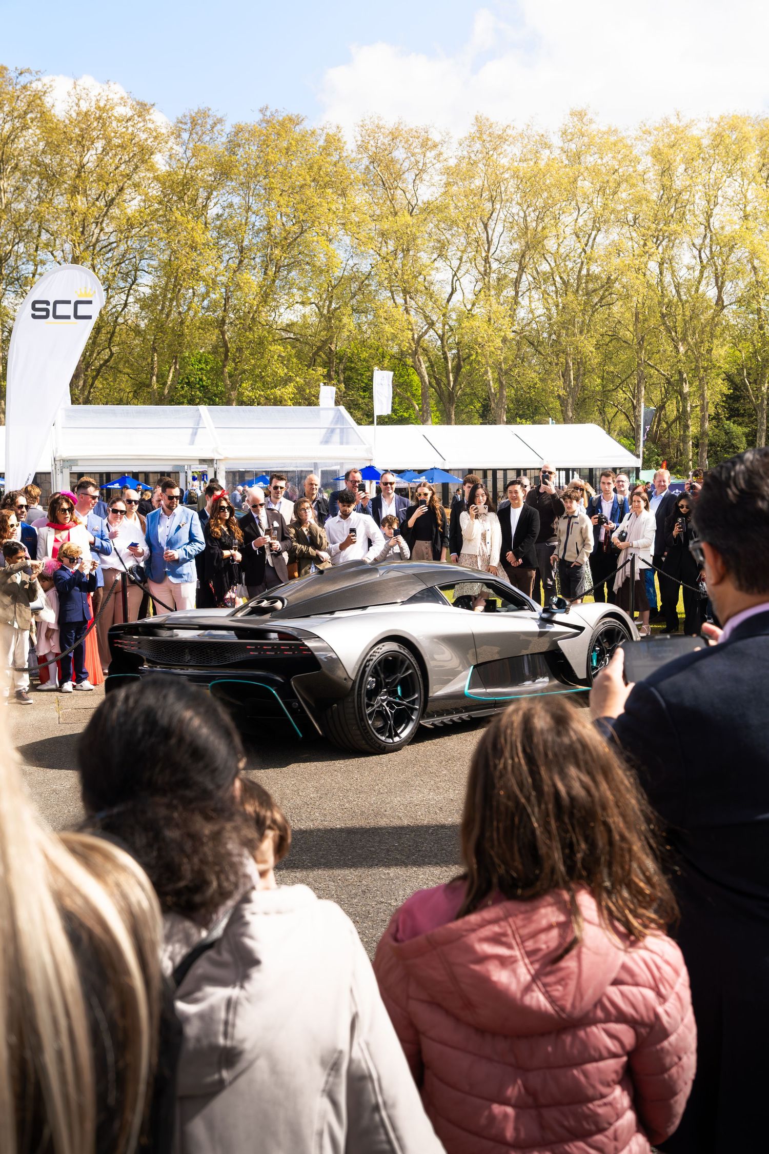 Crowd surrounds a sleek silver supercar at an outdoor SCC automotive show on a sunny day.