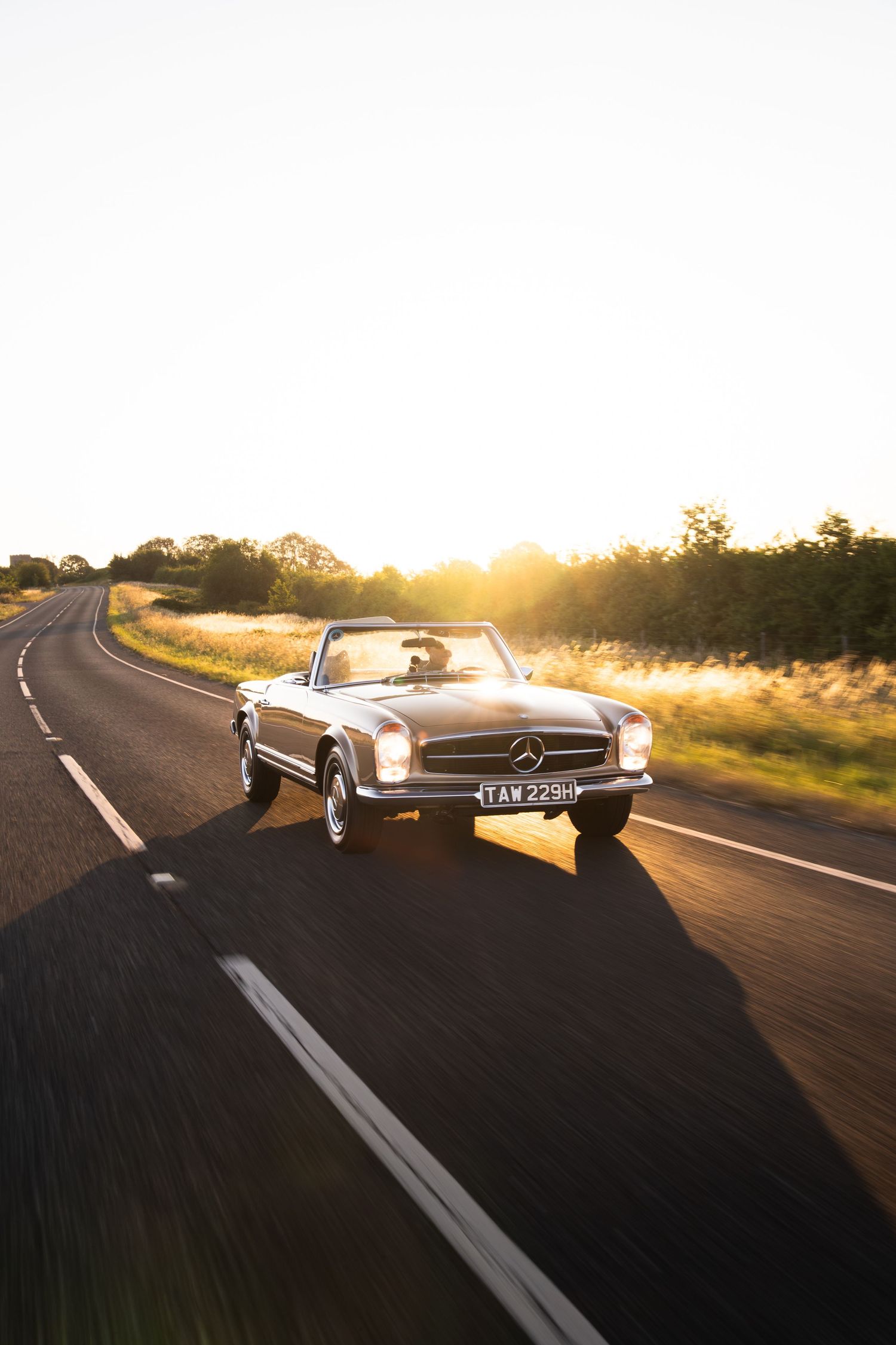 Classic Mercedes-Benz convertible cruising on a scenic road at golden hour sunset.