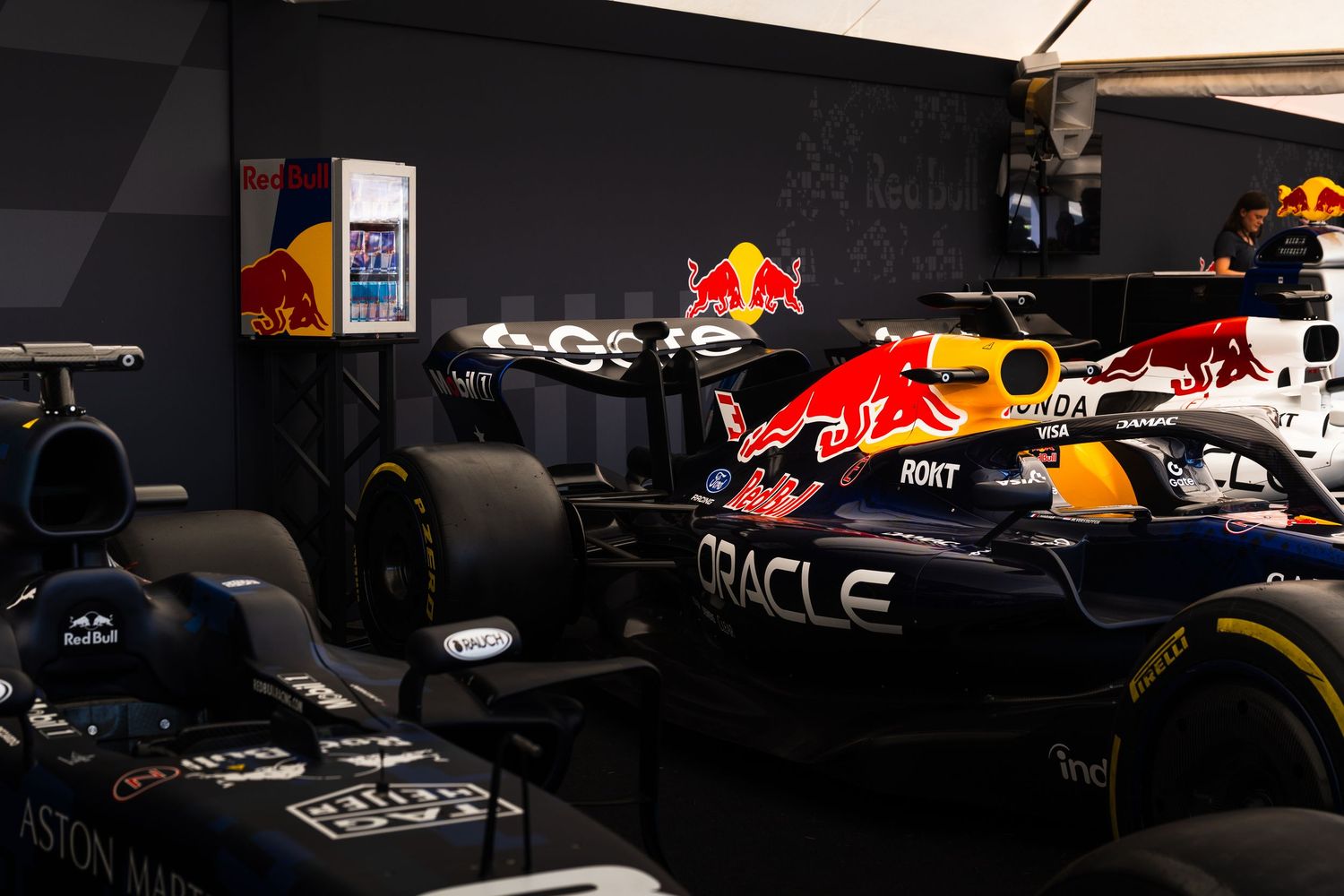 Red Bull Racing Oracle F1 car parked in the paddock garage alongside an Aston Martin Formula 1 car.