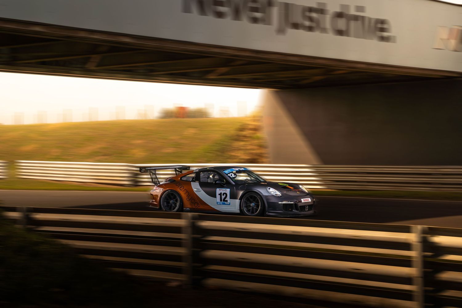 Racing car number 12 speeding on track at golden hour under bridge overpass.