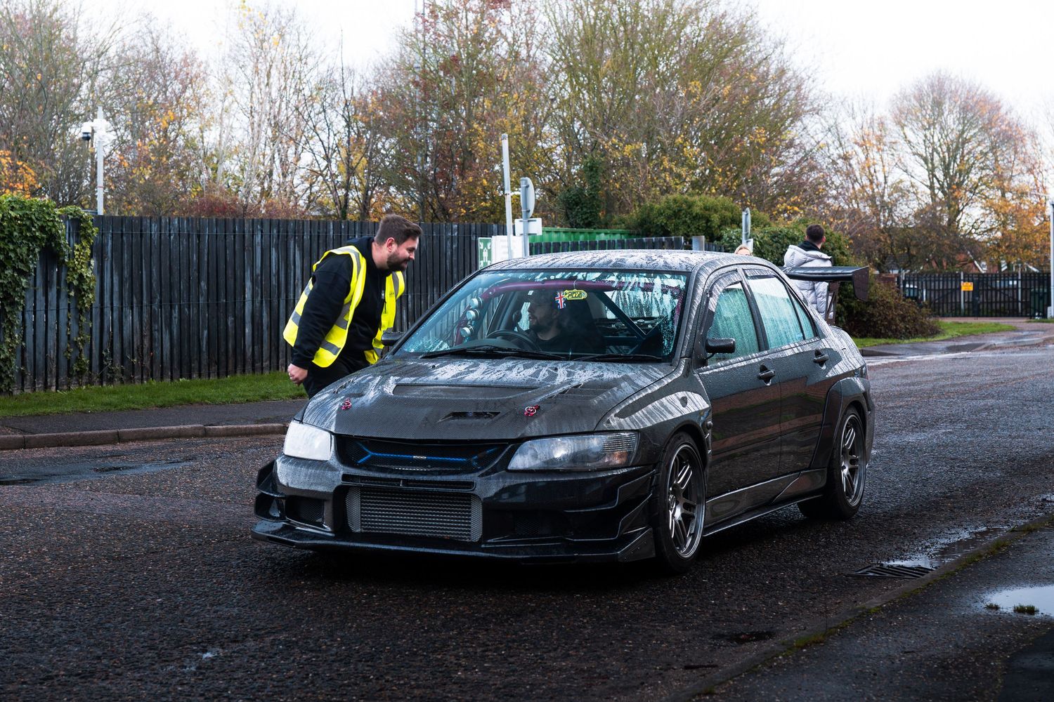A marshal in a hi-vis jacket inspects a dark modified Mitsubishi Evo at an outdoor event.