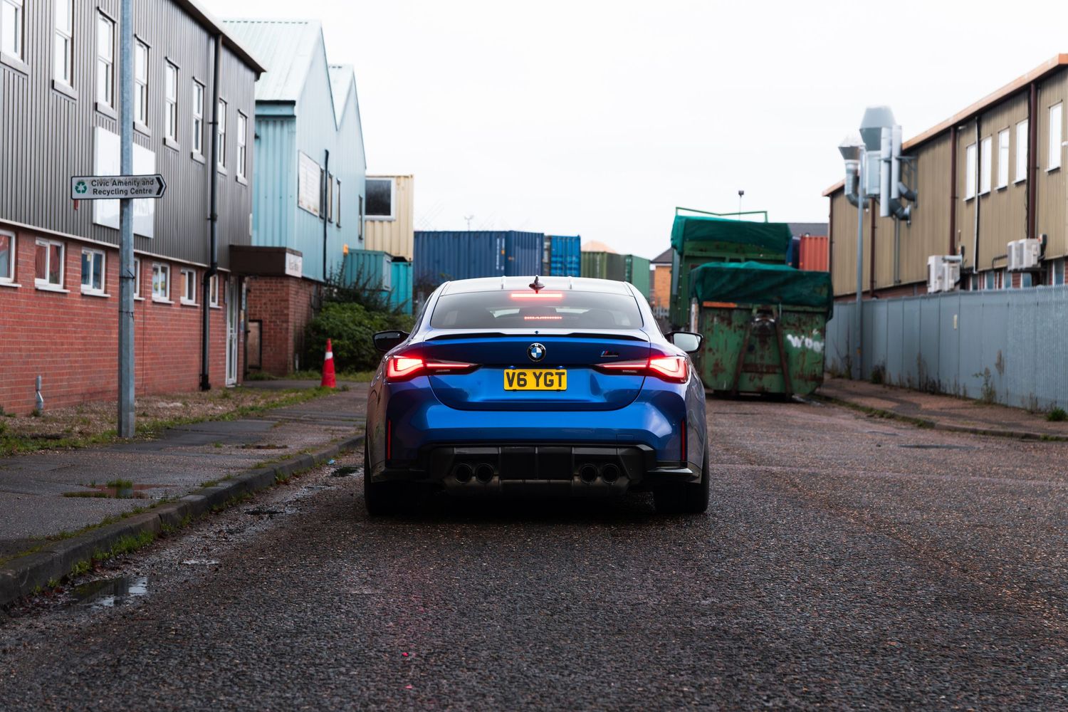 Blue BMW M4 rear view with active brake lights parked in UK industrial estate setting.