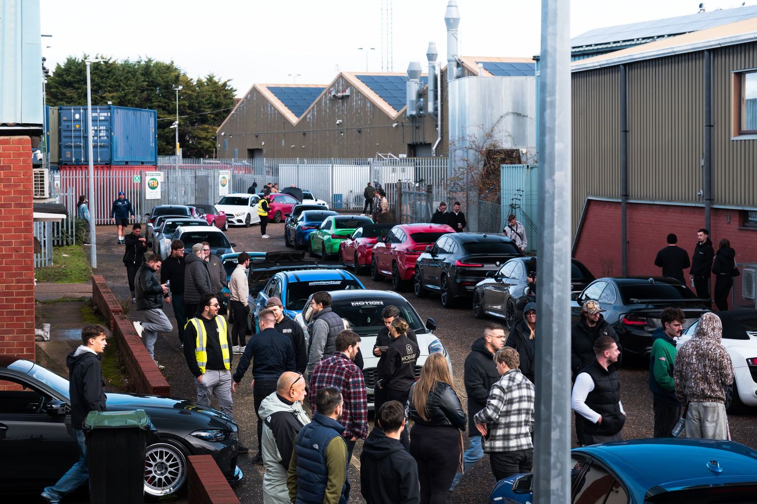 Large crowd of car enthusiasts gathered at an outdoor automotive meet in an industrial estate.