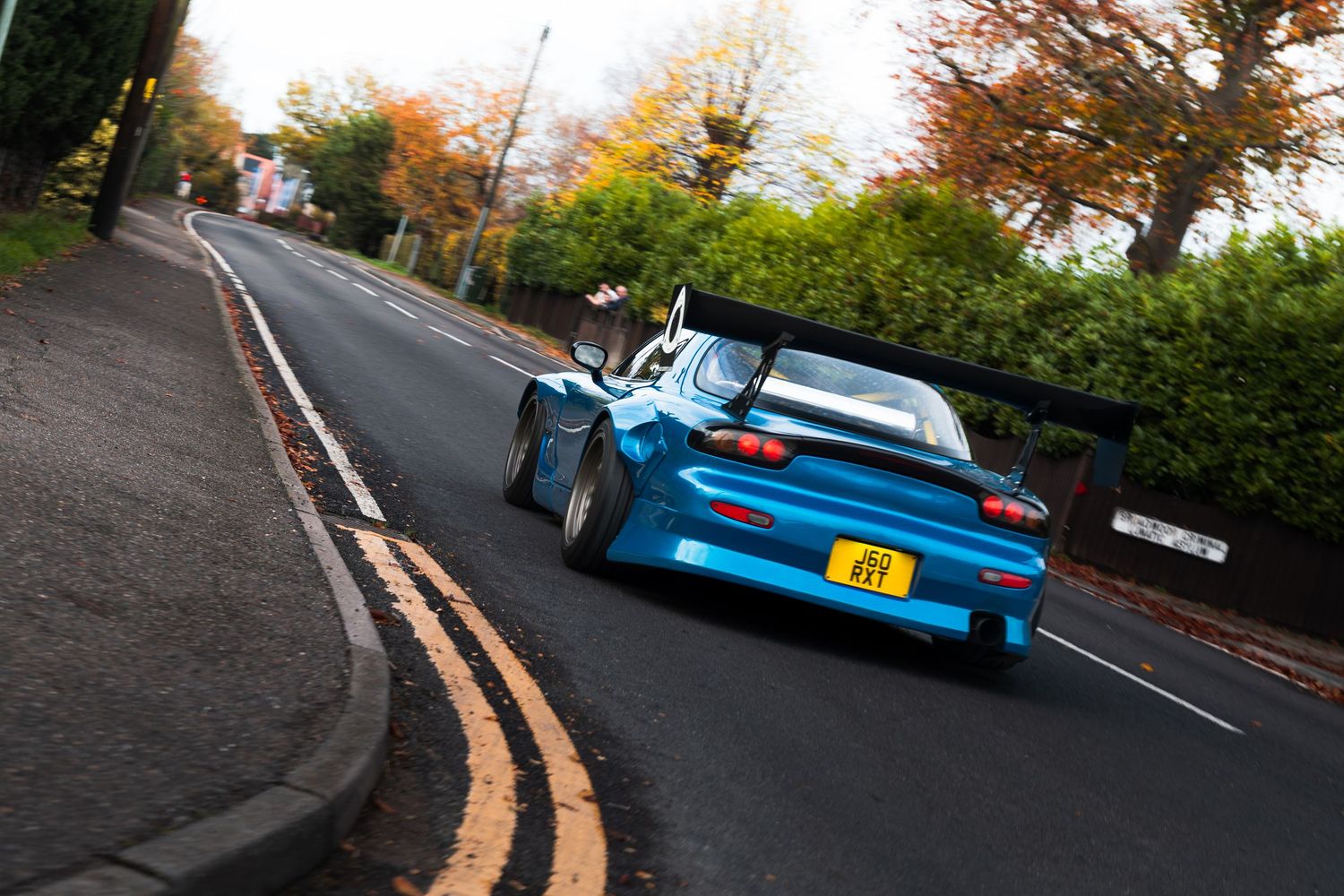Blue widebody Mazda RX-7 with large spoiler accelerating around a bend on a UK road.