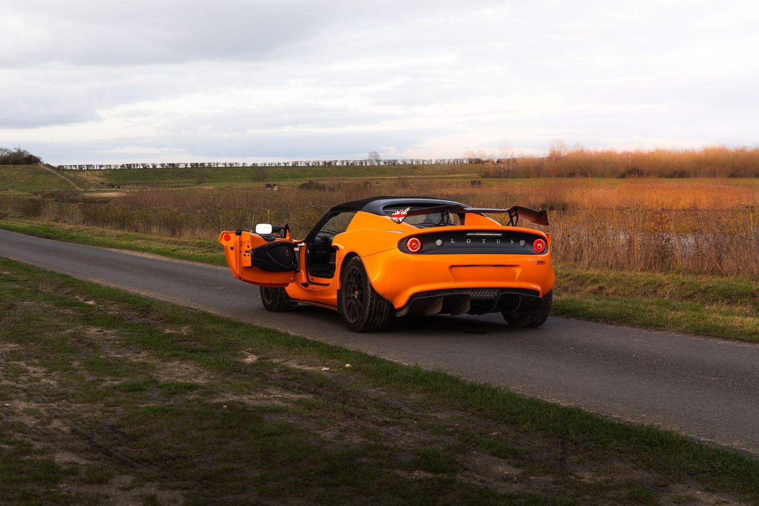 Orange Lotus Elise sports car driving on rural road with open doors.