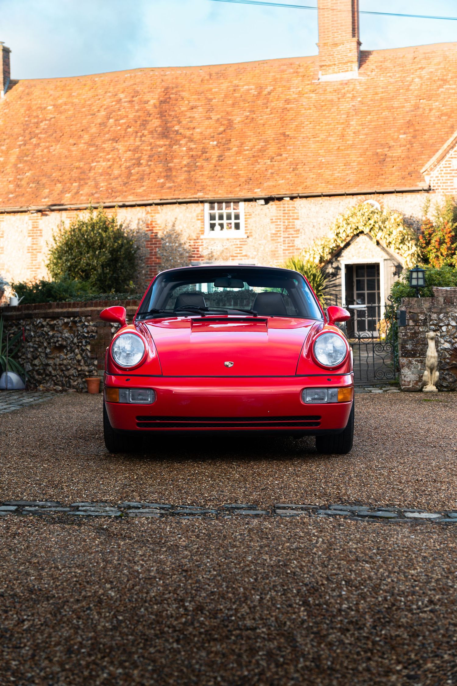 Front view of a red Porsche 911 Carrera 2 Targa parked on a gravel driveway before a brick farmhouse.