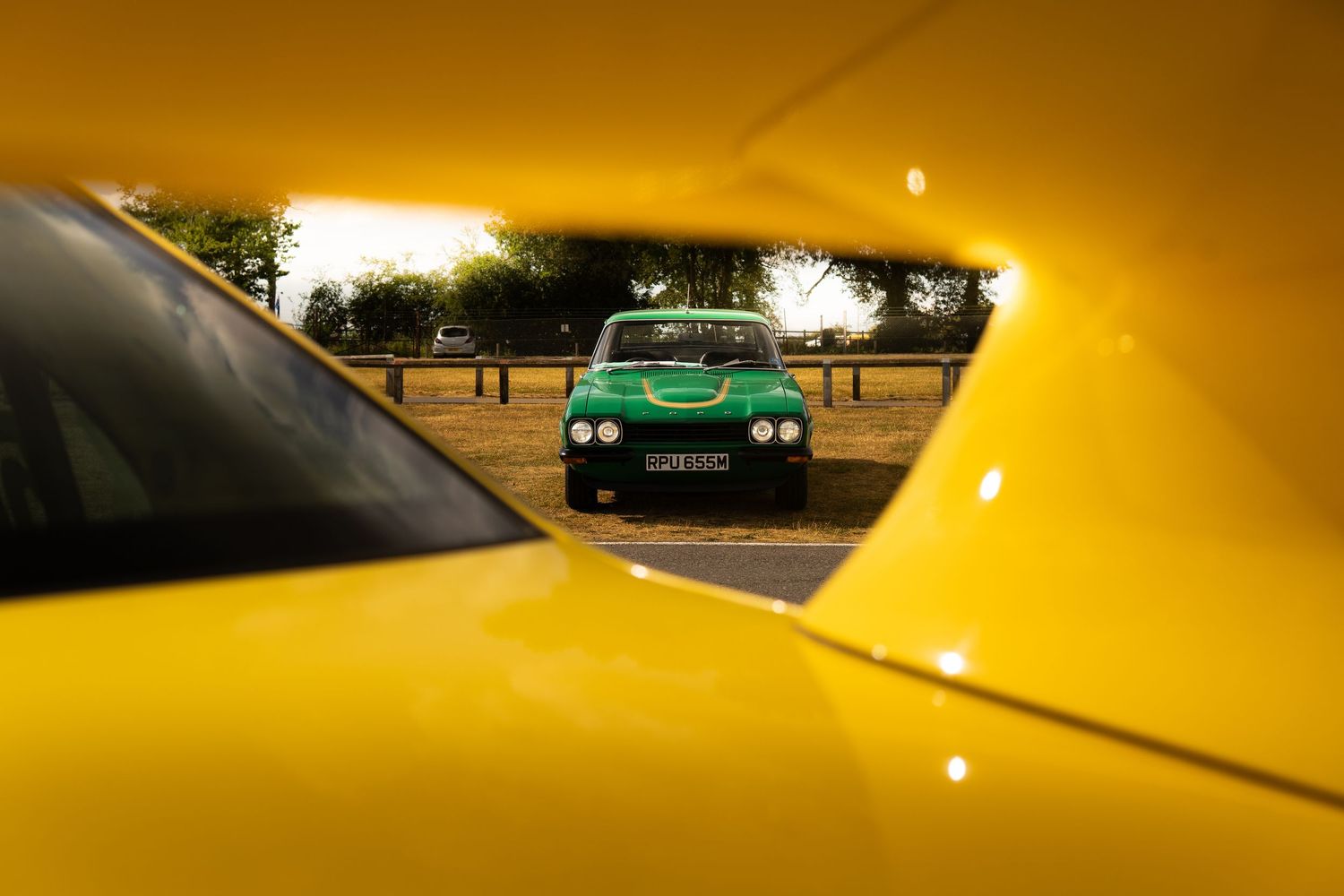 A green classic car viewed through the bodywork of a yellow car outdoors.