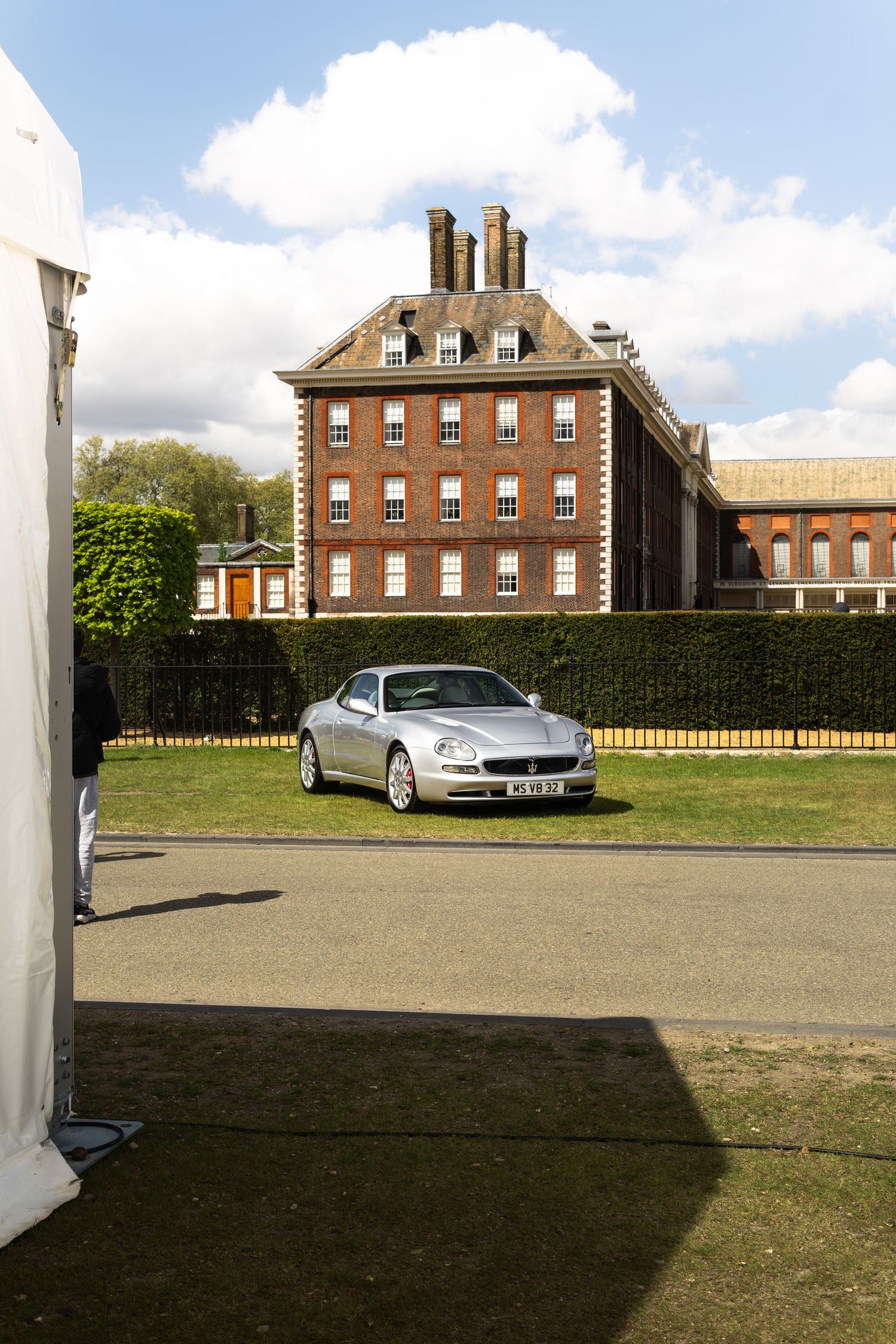 Silver Maserati coupe parked on grass in front of a classic red brick Georgian manor house exterior.