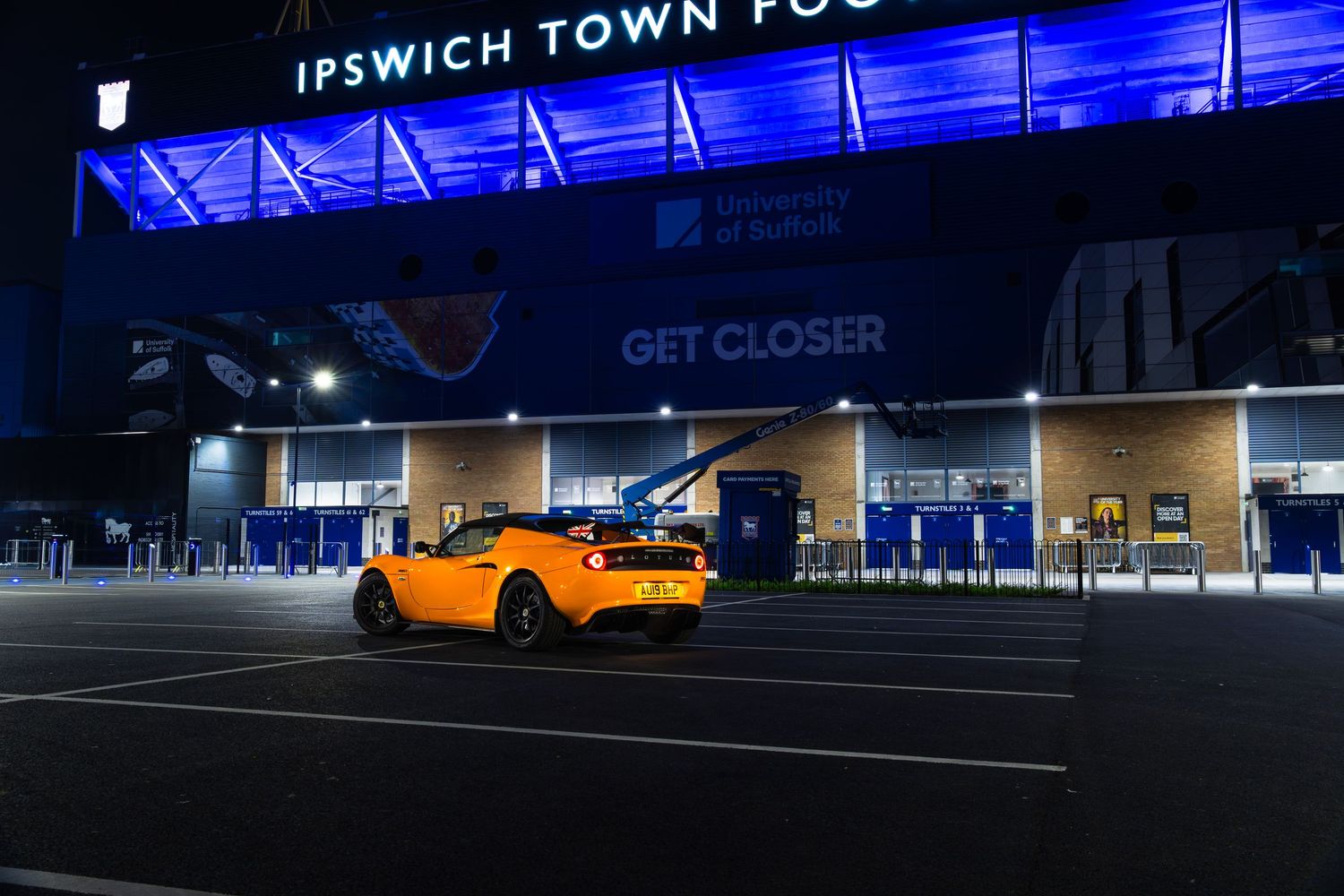 Orange Lotus Elise parked outside Ipswich Town Football Club stadium at night under blue floodlights.