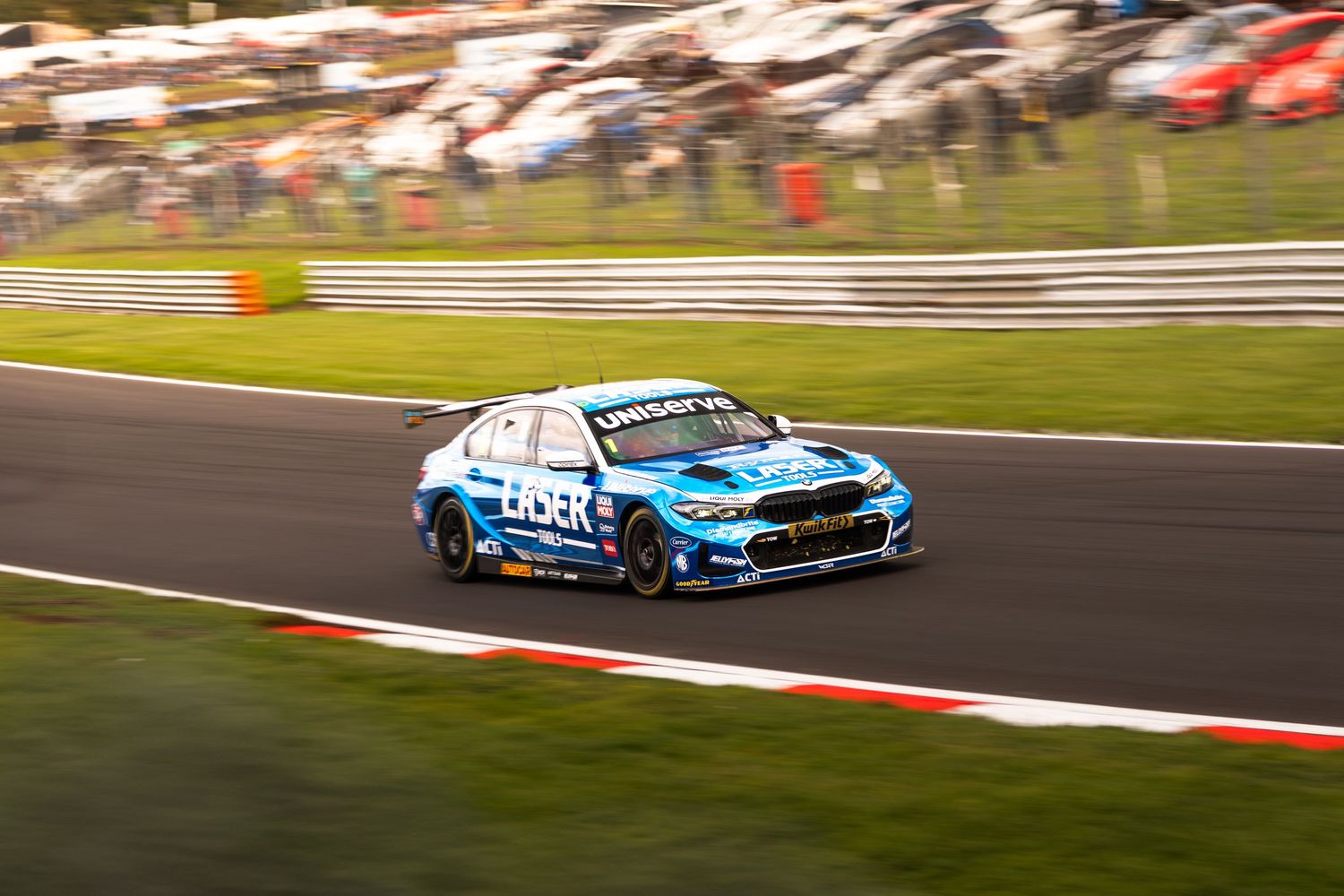 A blue BMW M2 competition BTCC race car navigates a corner at a motorsport circuit.
